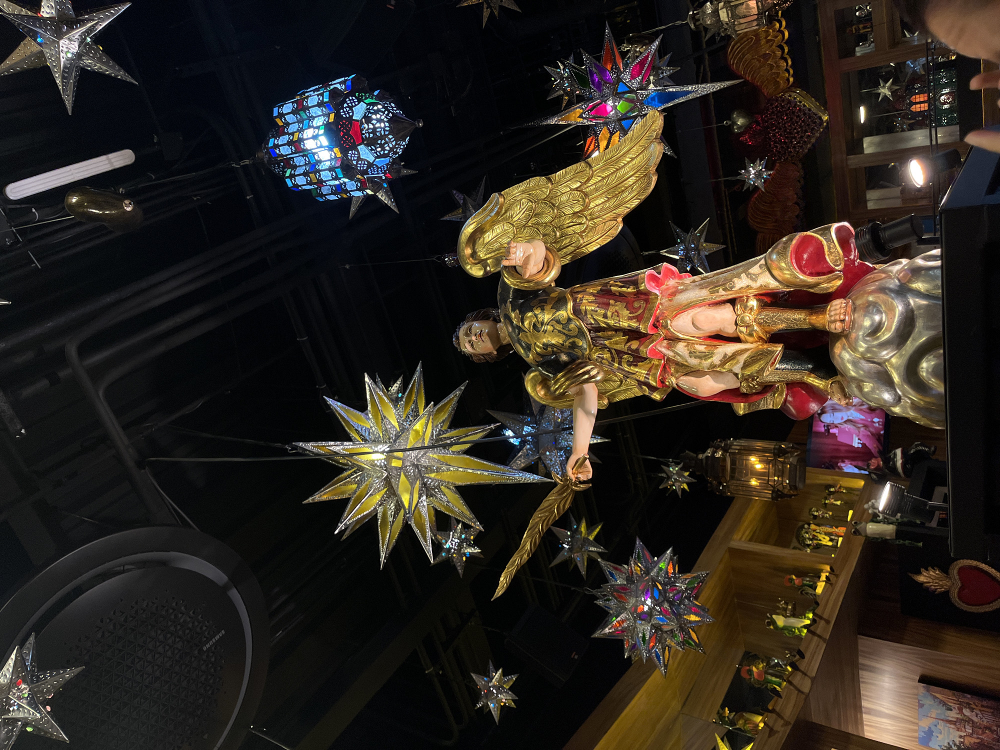 The image captures a low-angle view of a decorative space in Santa Lucía, Mexico. A golden statue of an angel with outstretched wings is centrally positioned. The statue is adorned with gold and red accents. The angel holds a gilded sword. Overhead, the ceiling is decorated with an array of hanging ornaments, including colorful star lanterns and a blue geometric light fixture. A large, circular Samsung air conditioning unit is visible in the bottom left. In the background to the right, a wooden shelf displays figurines. The overall lighting casts a warm, ambient glow.
