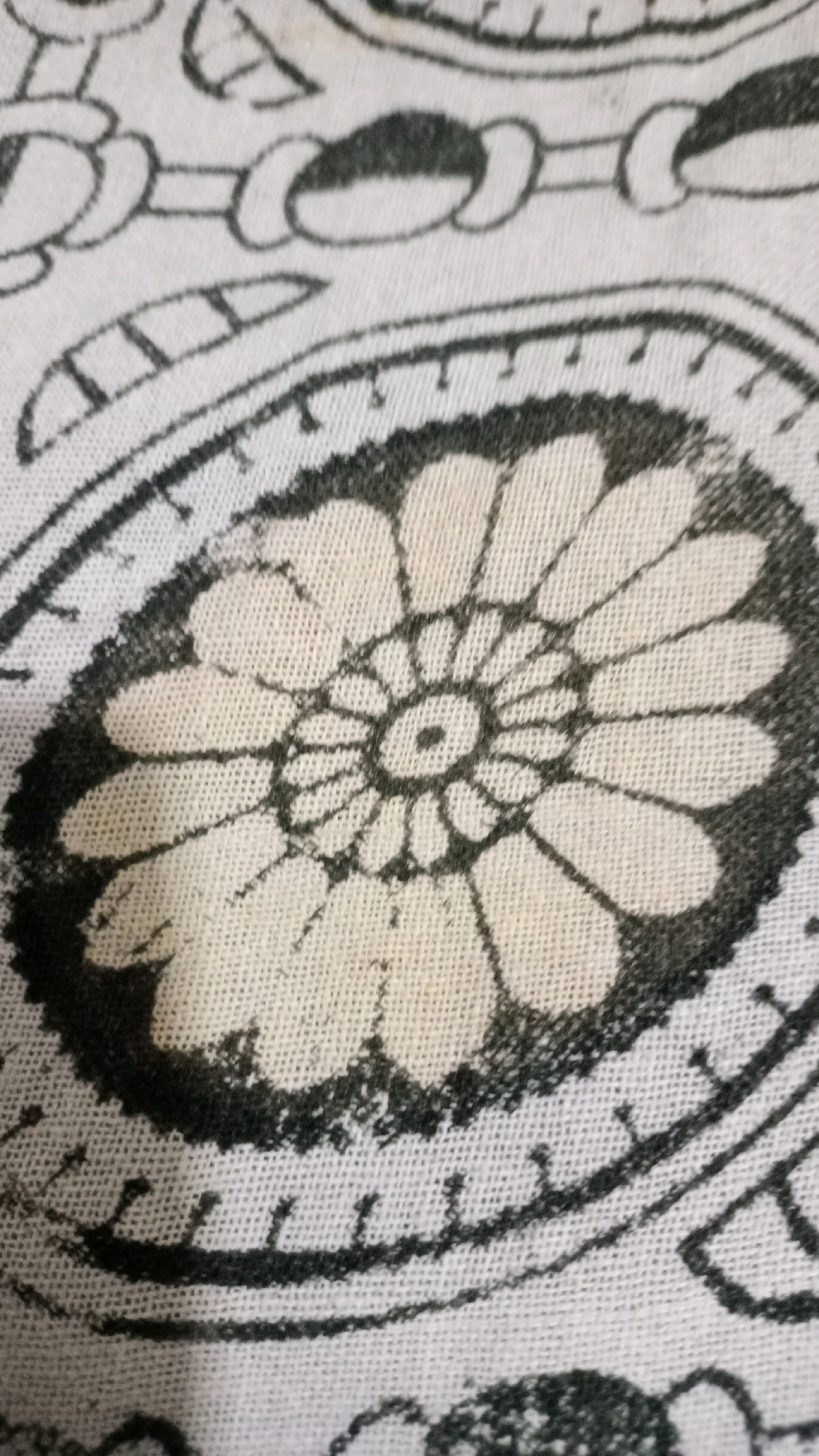 The image shows a close-up of a white textile with a black and cream floral pattern. The pattern features a central circular motif with radiating petals, surrounded by decorative elements resembling chains and stylized leaves. The textile appears to be made of cotton or a similar woven fabric, with visible threads creating a textured surface. The printing is somewhat faded and uneven, suggesting it might be a vintage or hand-printed item. There is no identifiable location in Deoghar, India, or any people or actions depicted in the image.