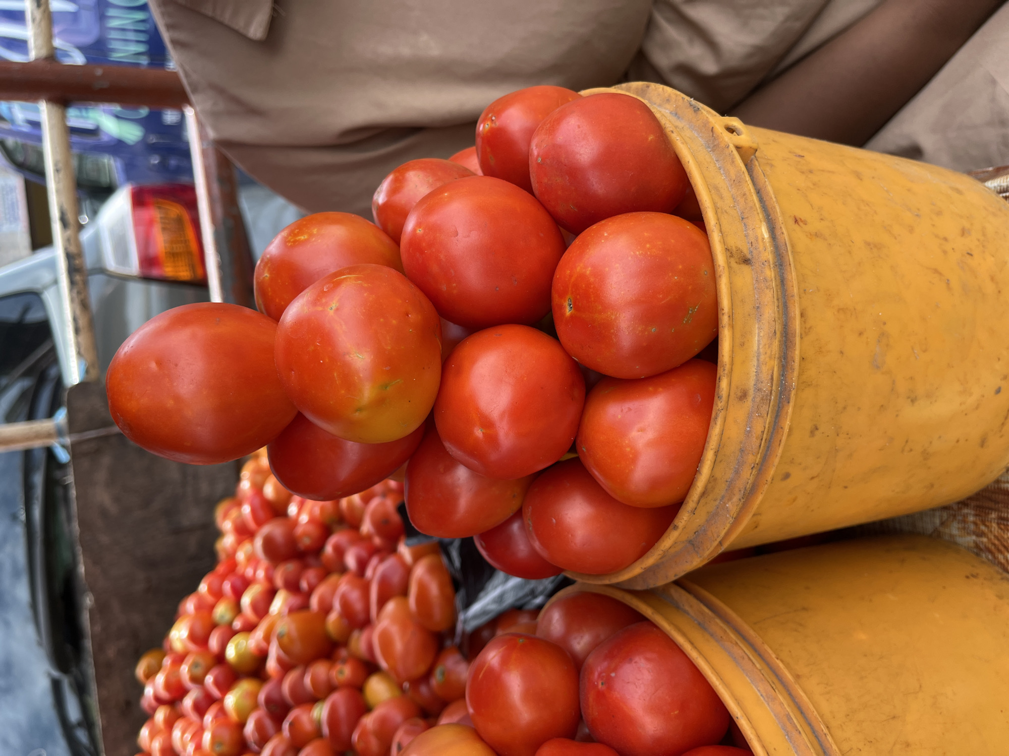The image shows two yellow plastic buckets overflowing with red tomatoes. One bucket is tilted slightly, with tomatoes spilling out. Below the buckets, there's a larger pile of tomatoes, some of which appear slightly less ripe, with yellowish hues. In the background, a person wearing a light brown garment is visible. Behind the tomatoes and buckets, elements of a vehicle and some metal structures are discernible. The scene is consistent with an outdoor market or roadside vendor setup in Lekki, Nigeria, where the sale of produce is common. The lighting suggests it's daytime.