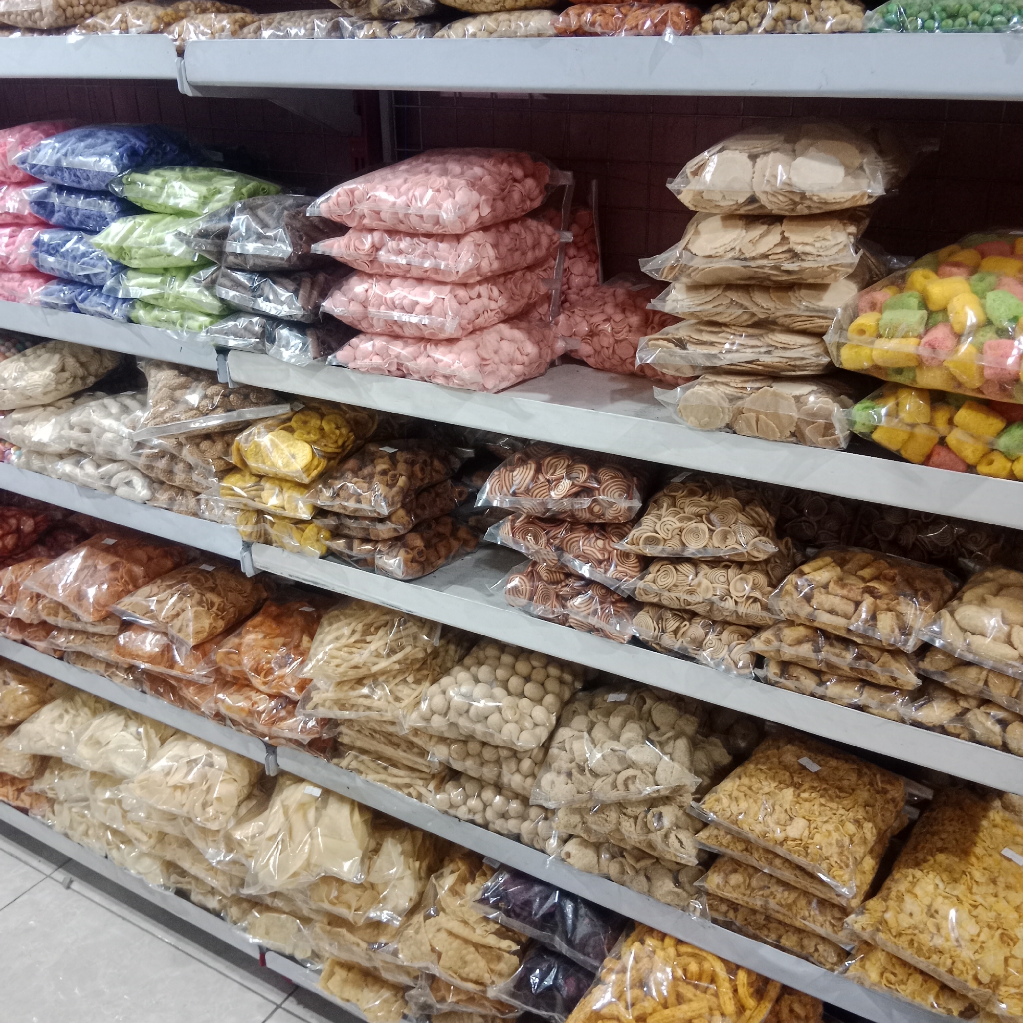 The image shows shelves in a store, possibly in Tegowanu Wetan, Indonesia, stocked with a variety of bagged snacks. The snacks are arranged in clear plastic bags and are stacked on multiple metal shelves. The types of snacks visible include colorful puffed snacks in shades of pink, green, yellow, and orange, as well as various types of chips and crackers that appear to be made from dough, some of which are light brown and circular or spiral-shaped, and others that are flat and yellowish. Some bags contain what look like small, round, light brown snacks, possibly nuts or puffed grains. The overall impression is of a retail environment displaying a wide selection of packaged snack foods. There are no people visible in the image. The lighting suggests an indoor setting.