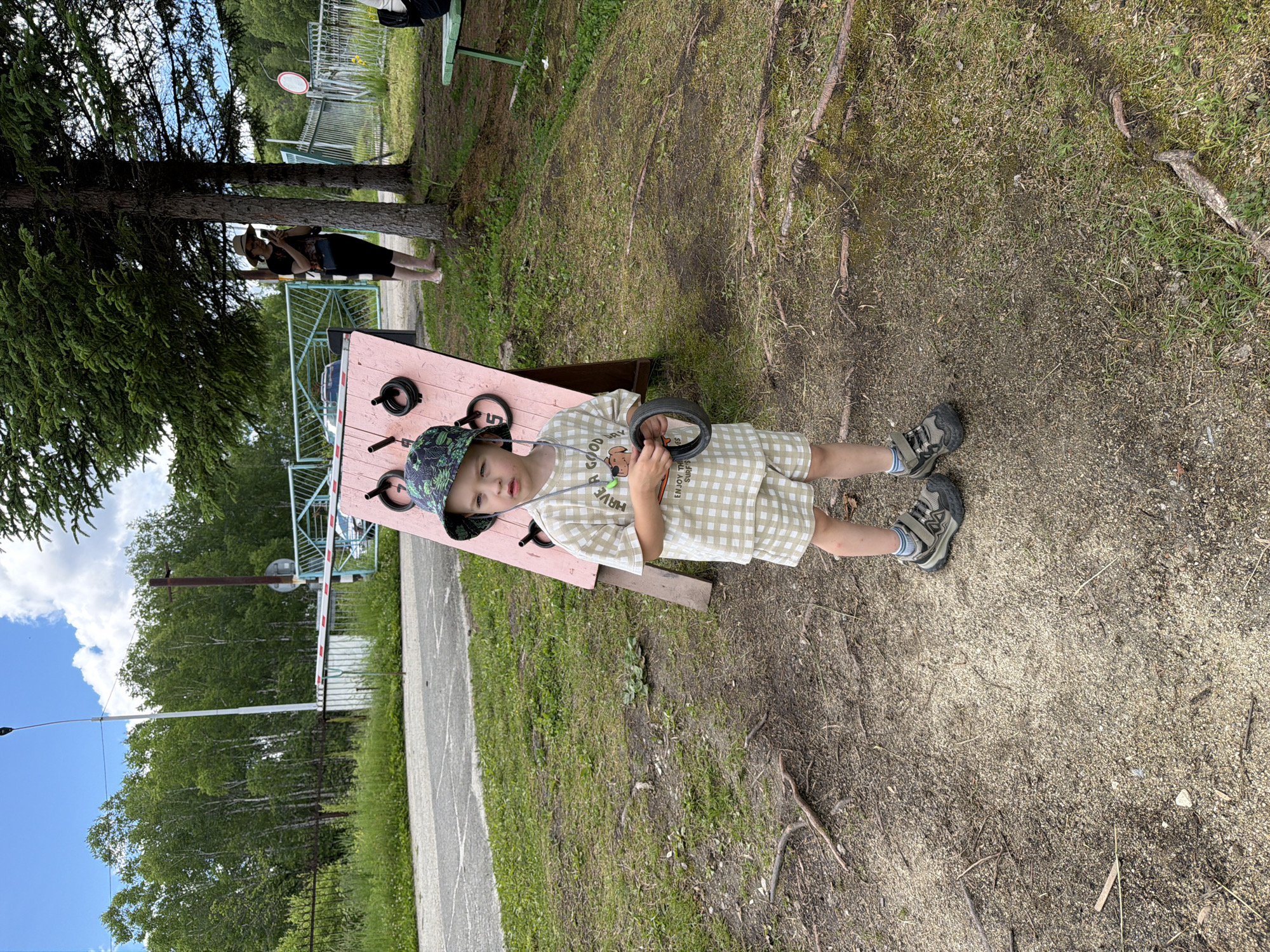A young child stands on a dirt path in what appears to be an outdoor recreational area in Kul'dur, Russia. The child is wearing a checkered outfit, a patterned bucket hat, and sneakers. They are holding a black ring in their right hand. Behind the child is a pink wooden structure with various circular openings and some black rings attached. In the background, trees line a fence, and a gate with a barrier is visible. A woman is standing under a tree to the right of the frame, looking at her phone. The ground is covered in dirt, small stones, and patches of grass. The sky is blue with scattered white clouds.