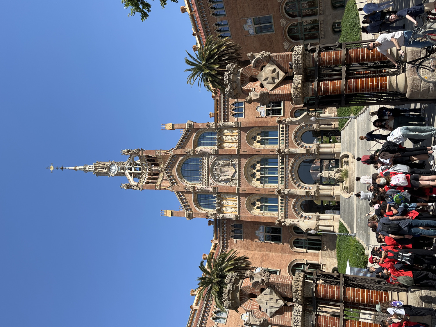 This media file presents a vibrant outdoor scene featuring the magnificent Modernisme architecture of the Hospital de Sant Pau in Barcelona, Spain. The primary subject is the grand, multi-story brick building, characterized by its intricate facades, arched windows, elaborate sculptural details, and a prominent central spire with a clock face. Palm trees flank the building, contributing to the Mediterranean setting.
In front of the building, a large group of people is gathered on a paved area, suggesting they are tourists or a school group. Many are looking towards the building, indicative of a guided tour or an appreciative visit. Some individuals are seen wearing red or white shirts, potentially signifying an organized group. Other people are casually walking or are with bicycles near the railings and stone pedestals that line the plaza.
The scene is set on a bright, clear day with a cloudless blue sky, suggesting midday or early afternoon. Strong sunlight illuminates the building's rich textures and details, casting defined shadows. The overall impression is one of active engagement with a significant cultural landmark. No discernible text is clearly visible on the building or among the crowd.