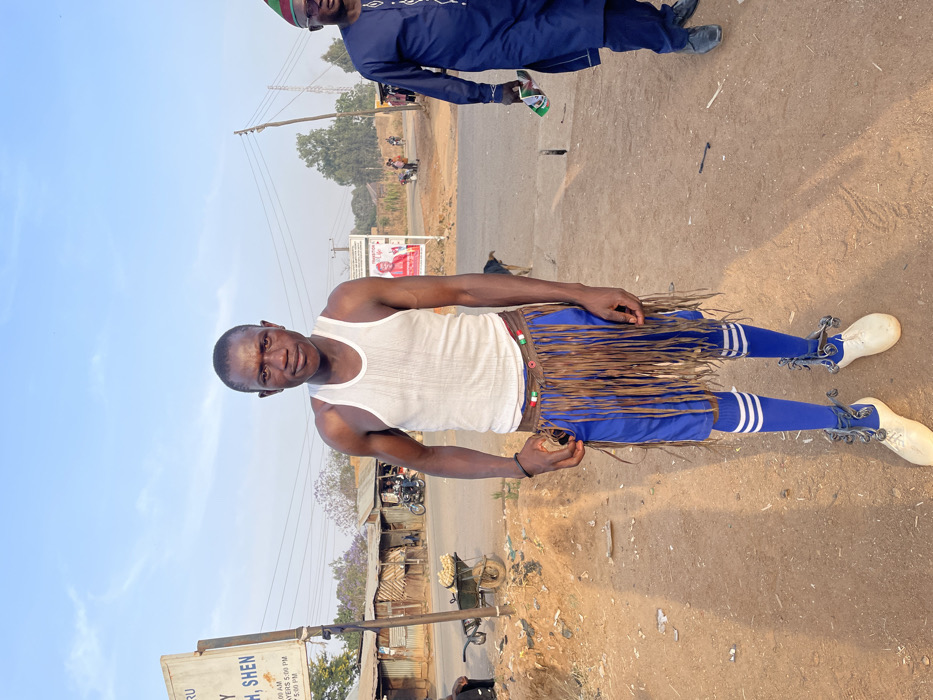 A man stands outdoors, wearing a white tank top, blue shorts and socks with white stripes. A fringed belt encircles his waist, and metal ankle bracelets adorn his feet. A blurred figure in a dark outfit stands slightly behind him. The background reveals a roadside scene with buildings, a sign, and vehicles, under a clear sky. The photo appears rotated, placing the horizon at a vertical angle.