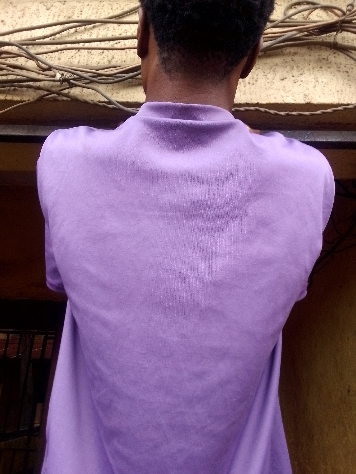 The image shows the back of a person wearing a purple shirt. The person is standing near a wall with exposed wires. The location appears to be an urban area, possibly outdoors. The city and state are unknown.