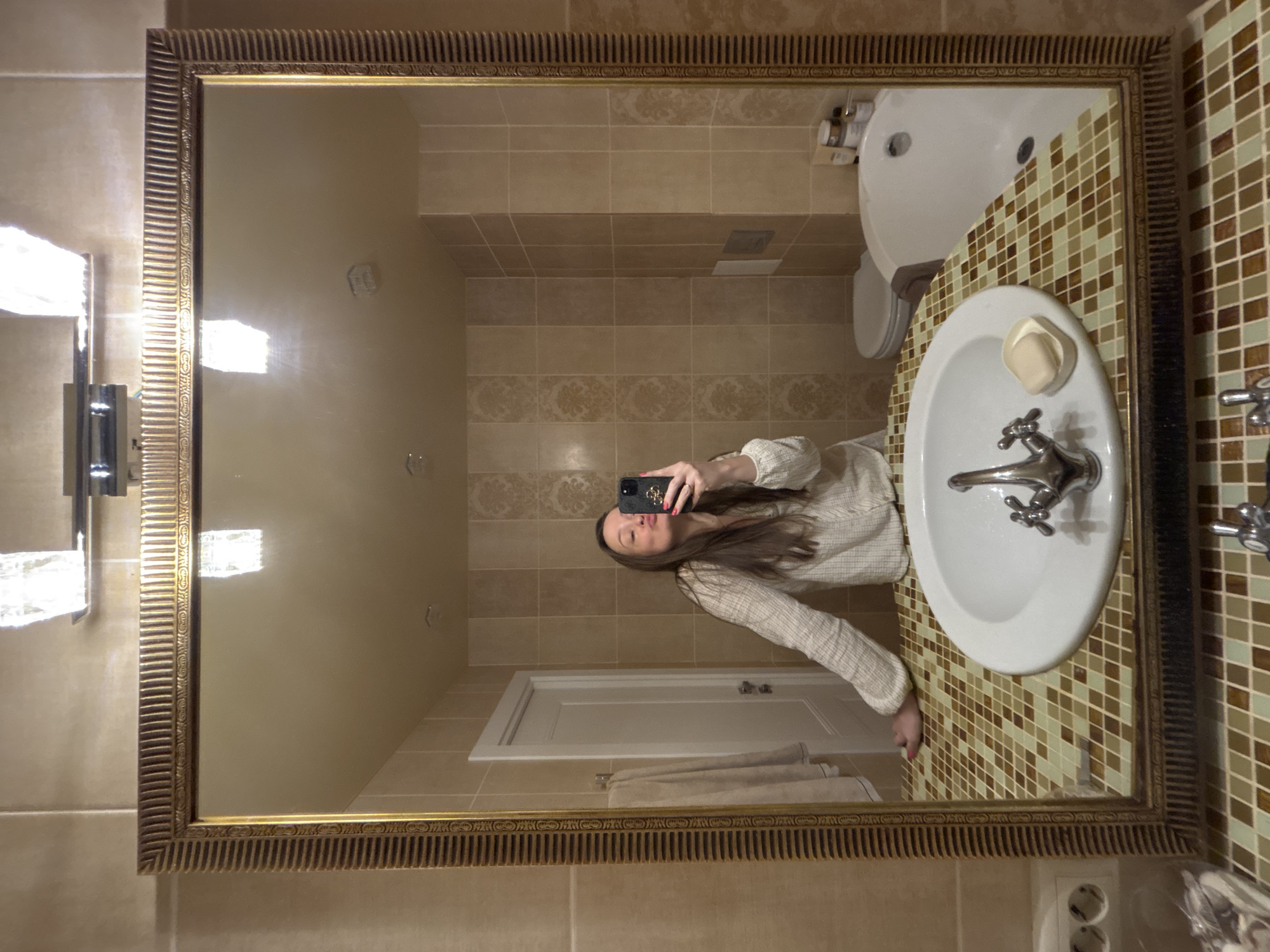 A young woman is reflected in a large, ornate-framed mirror in a bathroom, taking a selfie with her smartphone. The bathroom features tan tiled walls and floor, a white sink with chrome fixtures and a bar of soap, a toilet, and a towel rack with a towel. The sink's countertop is covered in multicolored mosaic tiles. In the mirror's reflection, a door is visible. The location is most likely Chaikovsky, Russia.