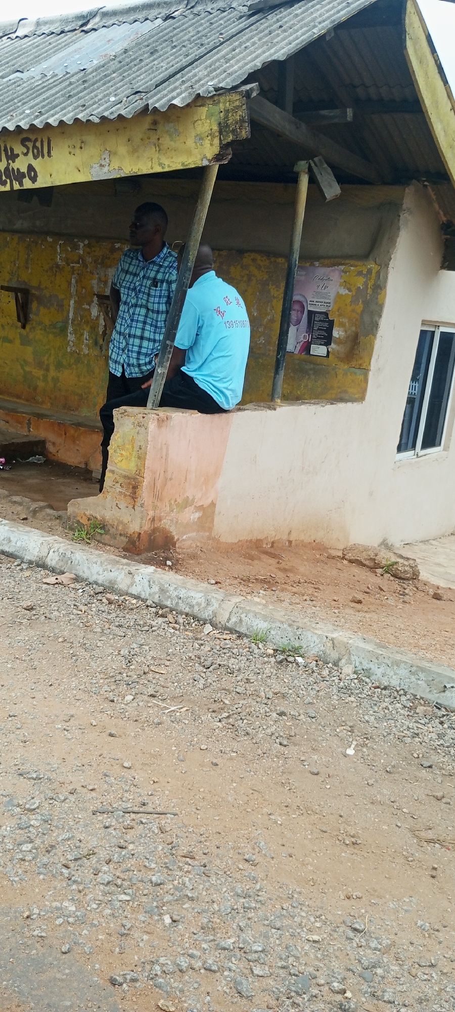The image shows a rustic outdoor scene in Adankolo, Nigeria, likely a bus stop or local gathering area. In the foreground, a gravelly dirt road with a concrete curb dominates the lower half of the frame. The ground is uneven and scattered with stones and debris.
Behind the curb, a weathered, makeshift shelter with a corrugated metal roof supported by wooden poles is visible. The walls of the shelter are peeling and painted in faded yellow and green. Two men are present. One man, wearing a blue and green plaid shirt and black pants, stands facing left, looking away from the camera. The other man, wearing a bright turquoise t-shirt with Chinese characters and a phone number printed on it, is seated on a low, concrete structure that appears to be part of the shelter's foundation. He is facing away from the camera.
A poster is affixed to the wall behind the seated man, featuring a portrait of an older man and text that appears to be an announcement or tribute. To the right of the shelter, a portion of a cream-colored building with a closed window is visible. The overall impression is one of a functional, community-oriented space with signs of age and wear.