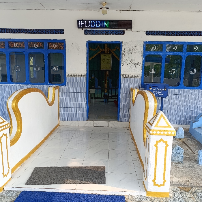 The image captures the entrance to a religious building, likely a mosque or Islamic prayer hall, characterized by its vibrant blue and white facade and distinct Islamic decorative elements. The building features a white exterior wall with a light blue and white checkered tile pattern along the lower sections, framing the doorway and windows.

A central open doorway leads into a darker interior, where a green curtain and a framed piece of text (likely religious scripture) are visible. Above the doorway, an LED display sign prominently shows the name "IFUDDIN" in glowing green and white letters. A simple light fixture is mounted on the ceiling above this sign.

Flanking the doorway are two windows with blue frames and multiple panes. Each pane contains a white decal featuring a crescent moon and star symbol at the top, and the Arabic word "الله" (Allah) below it.

In the foreground, a tiled pathway, light-colored and clean, leads up to the entrance. This path is bordered on both sides by low, decorative white walls with undulating, wave-like tops, highlighted with gold trim. These walls terminate in square, pillar-like structures also adorned with gold. A dark grey doormat is placed directly in front of the main entrance on the tiled floor. Partially visible at the bottom of the frame is a larger blue mat or carpet, extending outwards.

To the right of the pathway, beyond the low decorative wall, a blue box resembling a mailbox is visible. It has text on its side, which reads "Amal Jariyah," followed by "Pengurusan" and "Tanah Sara." This suggests it is a donation box for ongoing charity or for the management of the Sara land/property. Further to the right and slightly behind the mailbox, several small, light-colored stone markers or structures are visible, possibly indicating graves or decorative elements within a small adjacent area.

The scene appears well-lit, suggesting it is daytime. The overall impression is one of a well-kept and inviting religious site.