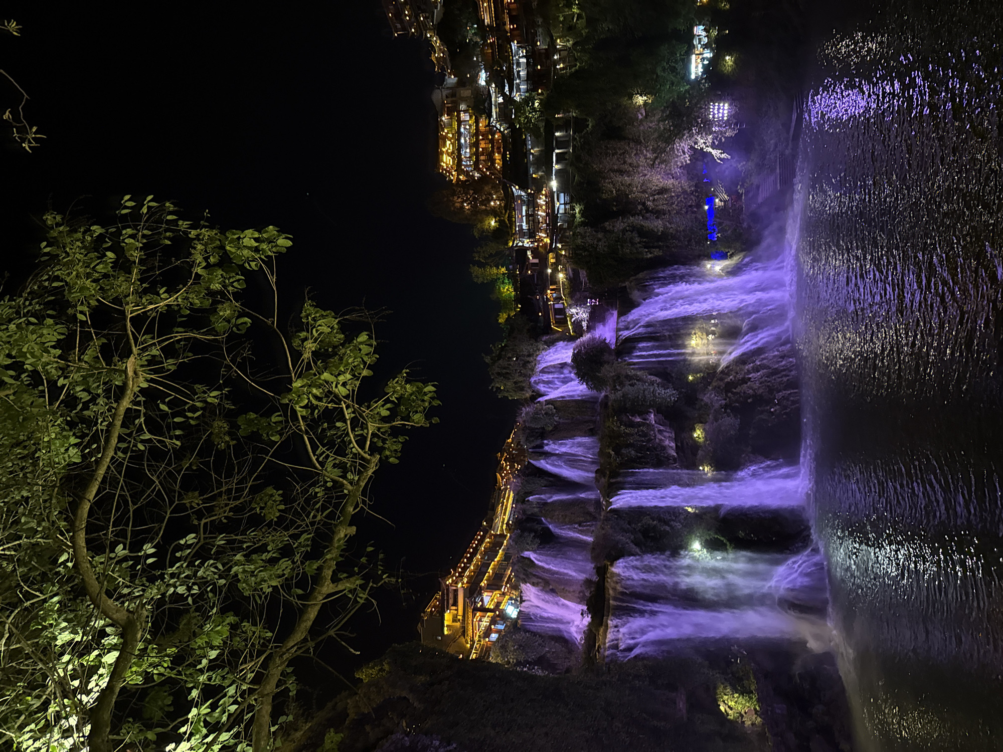At night, the ancient town of Furong in China is depicted with its picturesque cascading waterfalls illuminated with shades of purple. A tree with green leaves is prominent in the foreground, and the town's illuminated buildings are visible in the upper section of the image. The cascading waterfalls create a dramatic and captivating visual effect as they flow.