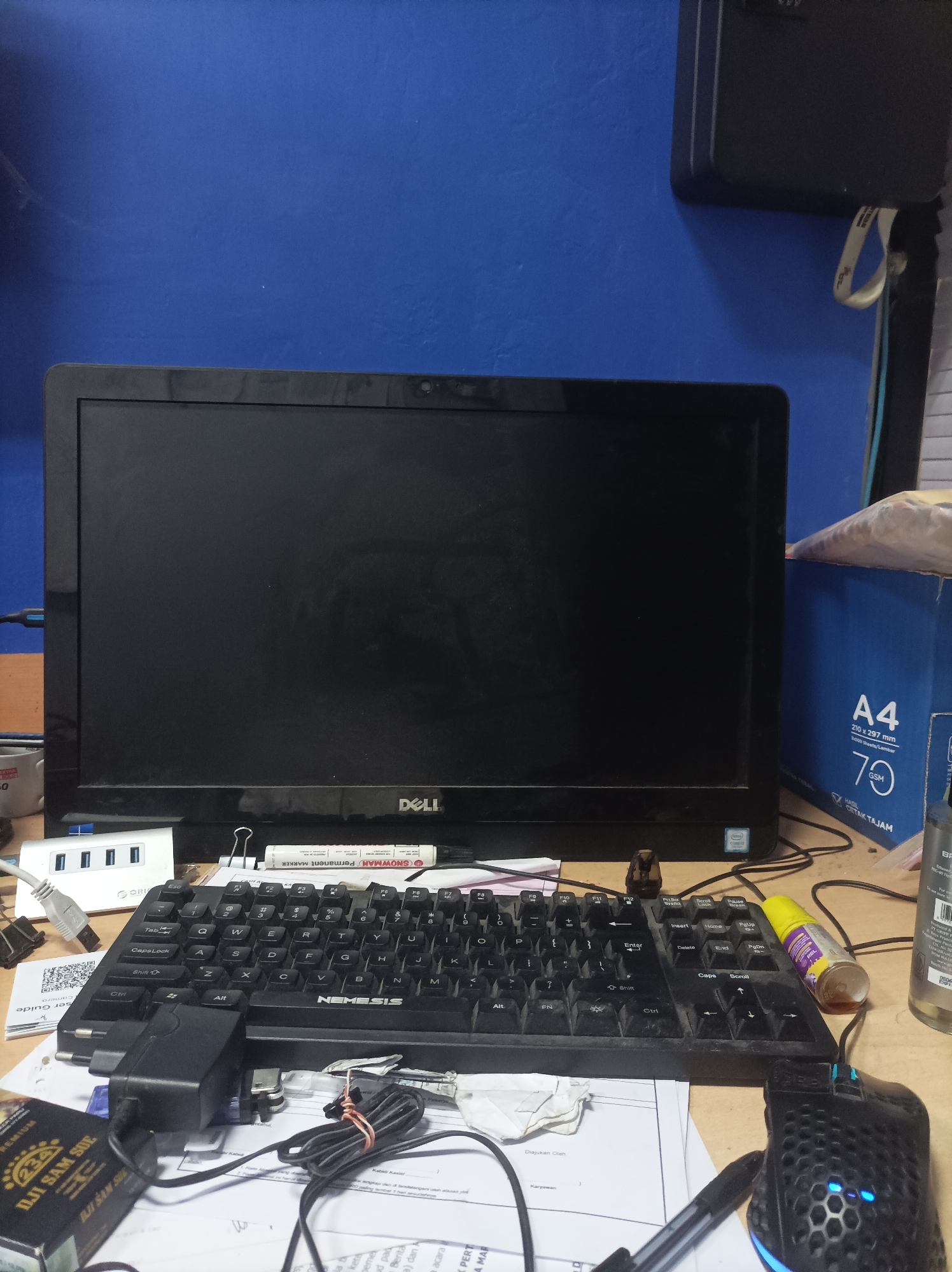 A close-up, eye-level shot captures a cluttered desk in what appears to be an office or workspace. The focal point is a black Dell monitor, with its screen turned off, displaying a reflective surface. In front of the monitor lies a black keyboard with Indonesian language labels on the keys. To the left of the keyboard, a white USB hub with four ports is visible, along with a connected white USB cable. A black power adapter with a coiled black cable is positioned in front of the keyboard. To the right of the keyboard, a black computer mouse with illuminated blue accents sits on a pile of papers.
Scattered across the desk are various objects, including a permanent marker, a roll of tape, and a small bottle with yellow liquid. Several documents and papers are present, some of which appear to be forms or receipts. In the background, a blue wall serves as the backdrop, and a large blue cardboard box, labeled "A4," is partially visible on the right side of the frame. A black laptop or external hard drive is perched on top of the monitor. The overall impression is one of a functional but disorganized workspace. The image is well-lit, with no strong shadows indicating artificial light. The composition is tightly framed, emphasizing the immediate desk area.