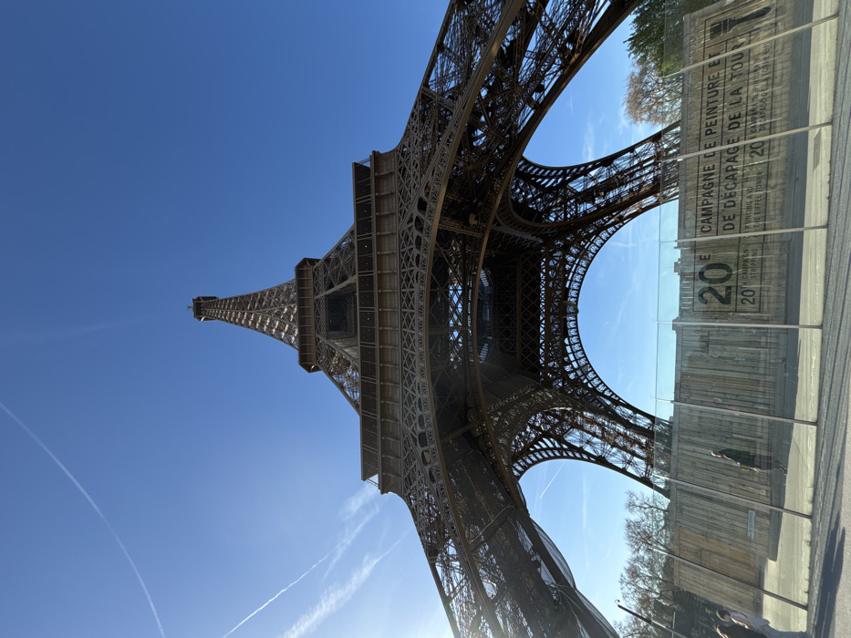 The image displays a low-angle view of the Eiffel Tower in Paris, France, against a clear, bright blue sky containing several faint white contrails. The tower's intricate iron lattice structure is the dominant subject, extending upwards from the base towards the zenith. Sunlight emanates from the upper left, casting some lens flare.
At the base of the tower, a clear barrier, likely constructed of glass or transparent plastic panels, runs horizontally across the foreground. Behind this barrier, a wooden hoarding or fence is erected. Prominently displayed on the hoarding are large letters and numbers, which, despite reflections on the clear barrier, are decipherable as "20E CAMPAGNE DE PEINTURE ET DE DECAPAGE DE LA TOUR" (20th painting and stripping campaign of the tower) in French and "20TH CAMPAIGN OF STRIPPING AND PAINTING OF THE EIFFEL TOWER" in English, accompanied by a stylized paintbrush icon.
To the far left of the frame, behind the clear barrier and in front of the wooden hoarding, two individuals are partially visible, one wearing a light-colored garment and the other a darker one. Near the center-left, a solitary individual dressed in dark clothing is walking on a paved surface behind the clear barrier, adjacent to the wooden hoarding. Deciduous trees with bare branches are visible beyond the hoarding on both the left and right sides of the scene.