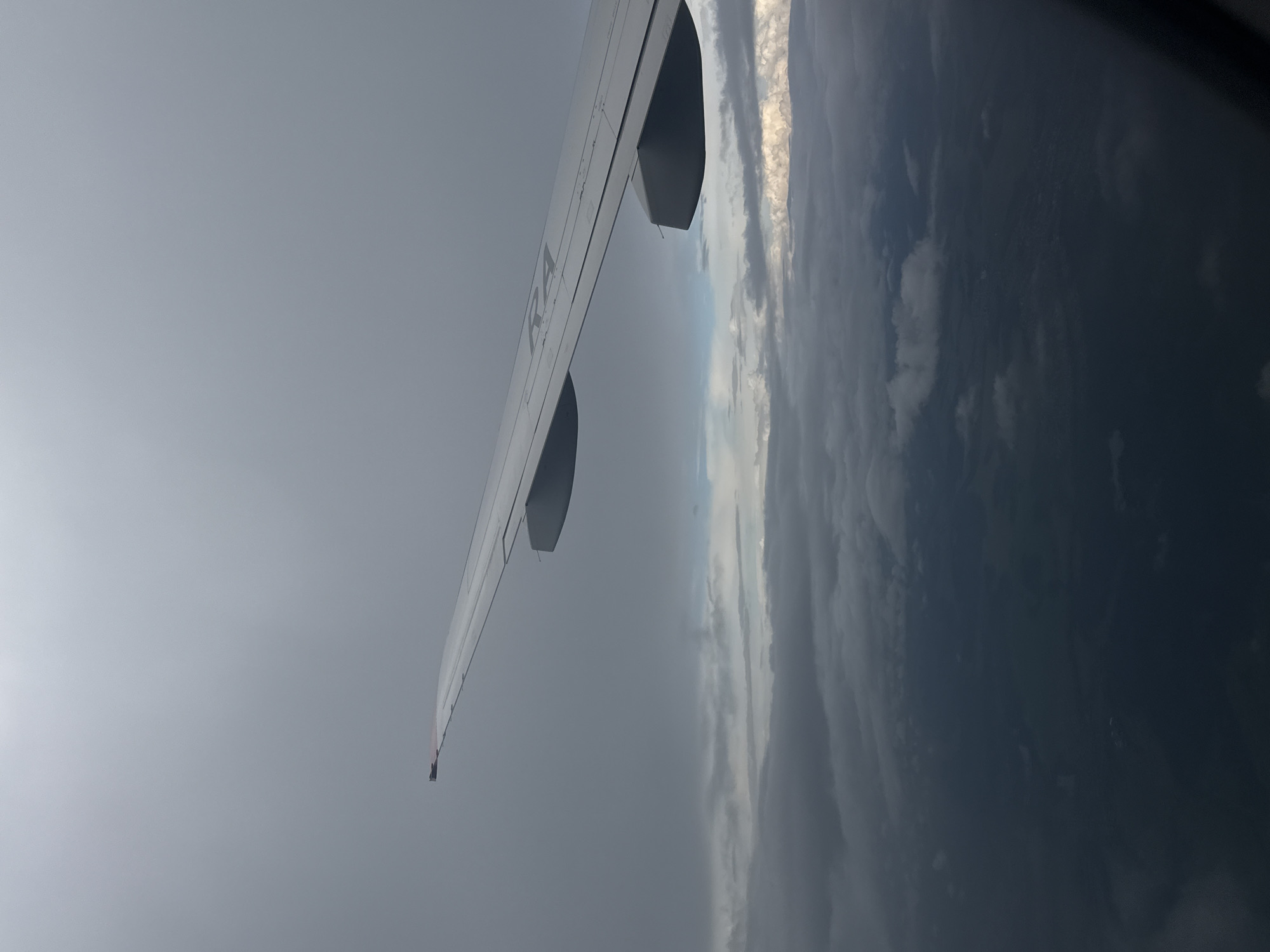 An aerial view from an airplane window shows a wing in the foreground, angled upwards and to the right. The wing is white with some markings, including what appears to be "RA" visible on its surface. Below the wing, a layer of dark, heavy clouds obscures the ground. Glimmers of lighter clouds are visible between the darker ones, suggesting a broken cloud layer. The ground below is largely obscured by the clouds, appearing as dark, textured shapes that could be fields or forests. The sky above the wing is overcast and grey. The image does not contain any information that would allow for its location to be identified as Staroe Mart'yanovo, Russia.