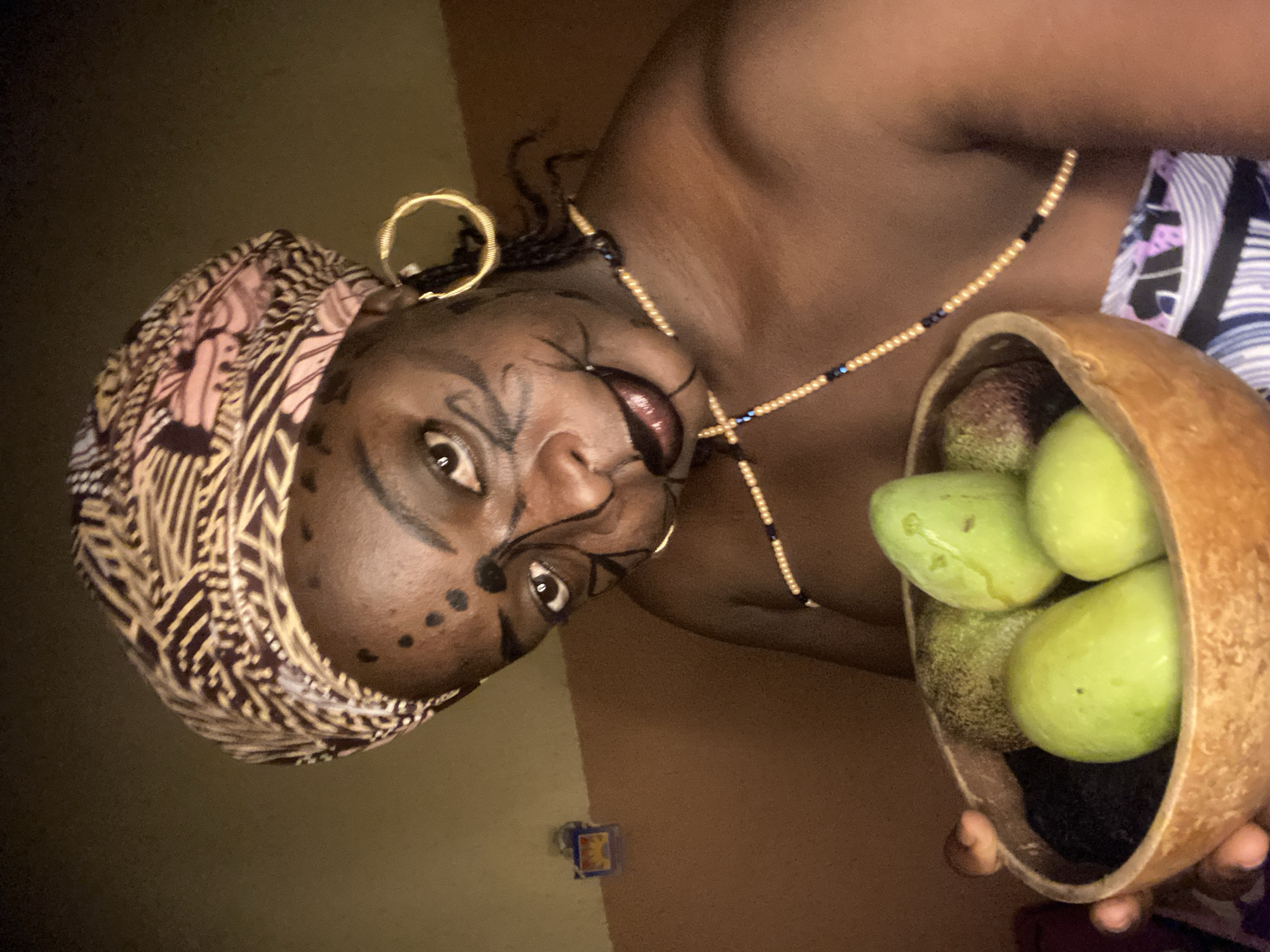 The image displays a person, likely a woman, with dark skin, adorned in striking cultural or artistic attire. Her head is covered with a patterned headwrap featuring intricate designs in shades of brown, black, and beige. A prominent gold-colored ear cuff is worn on her right ear, and a beaded necklace, consisting of gold/beige beads interspersed with a few black/blue beads, adorns her neck.
Her face is painted with elaborate black lines and dots, creating a stylized, mask-like design around her eyes and cheeks. Her lips are colored with a dark reddish-brown hue. She is holding a large, rustic bowl, appearing to be made from a dried gourd, which contains several unripe green fruits, resembling mangoes or avocados. The fruits vary slightly in size and texture.
The person's gaze is directed towards the camera, conveying a composed and deliberate expression. The setting appears to be indoors, against a plain wall with varying brown and beige tones, suggesting a room with potentially low or indirect lighting. A small, blurry, colorful object is visible on the lower wall to the left. The overall scene evokes a sense of cultural presentation, artistic expression, or a ritualistic portrayal, set within an interior environment in Maigizo, Nigeria. No discernible text is visible in the image.