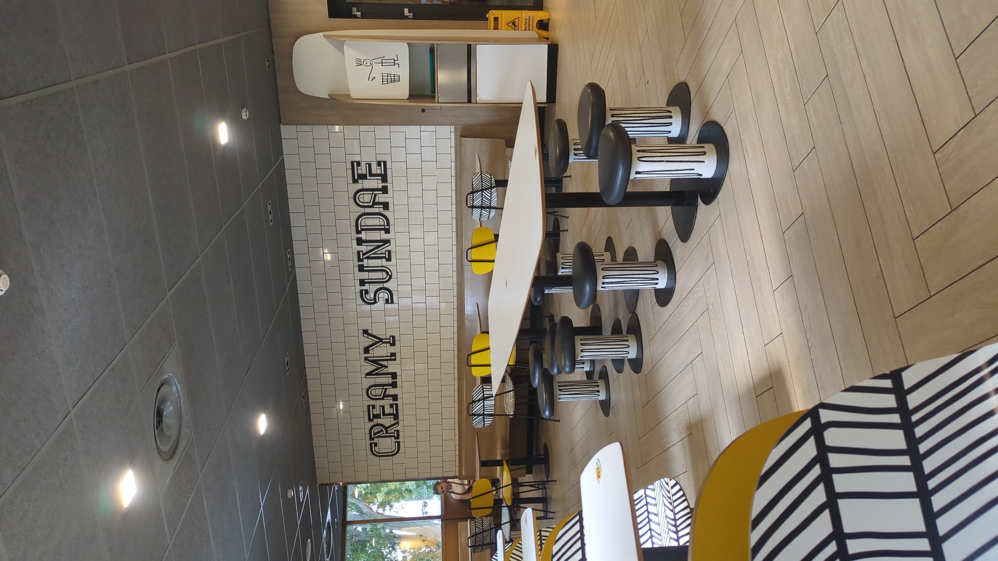The image depicts the interior of a fast-food restaurant, likely a McDonald's, given the decor and the "CREAMY SUNDAE" sign on the tiled wall. The scene is brightly lit, with a clean and modern aesthetic. The floor is made of light-colored wooden planks laid in a herringbone pattern.
In the foreground, a long, white, rectangular table is set with several black and white striped cylindrical stools. The stools have black circular bases and black padded seats. To the left and right of this central table, other tables and seating areas are visible. Some tables have yellow chairs with black and white patterned backs, while others have similar black and white patterned chairs.
A person, likely a customer, is visible in the background, sitting at a table near a window. The window looks out onto trees and foliage, suggesting an outdoor area.
On the right side of the image, there is a tall, white dispenser unit with a graphic of a person throwing trash into a bin. Below this, there is a yellow "wet floor" sign.
The ceiling is composed of grey acoustic tiles with embedded square lights. The overall impression is a well-maintained and inviting dining space. There is no indication that this scene is located in Chișinău, Moldova.