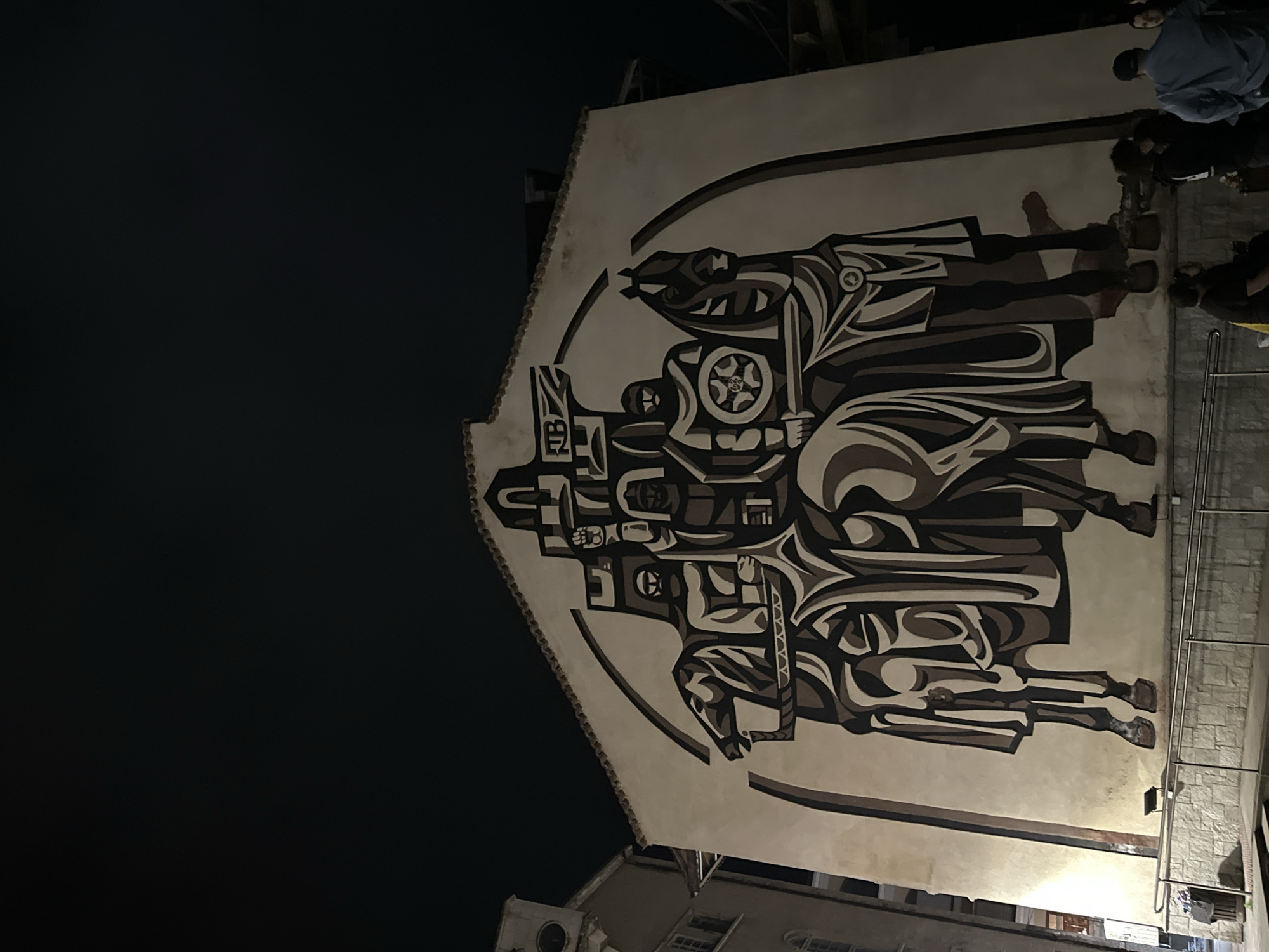 This image shows a mural on a building in Veliko Tarnovo, Bulgaria. The mural features stylized, abstract figures in shades of black, white, and gray, depicting a historical scene or figures. The mural is enclosed in a curved border. The sky is dark and the scene is lit by artificial light. Several people stand nearby, observing the mural. The surrounding architecture includes other buildings and a metal railing.