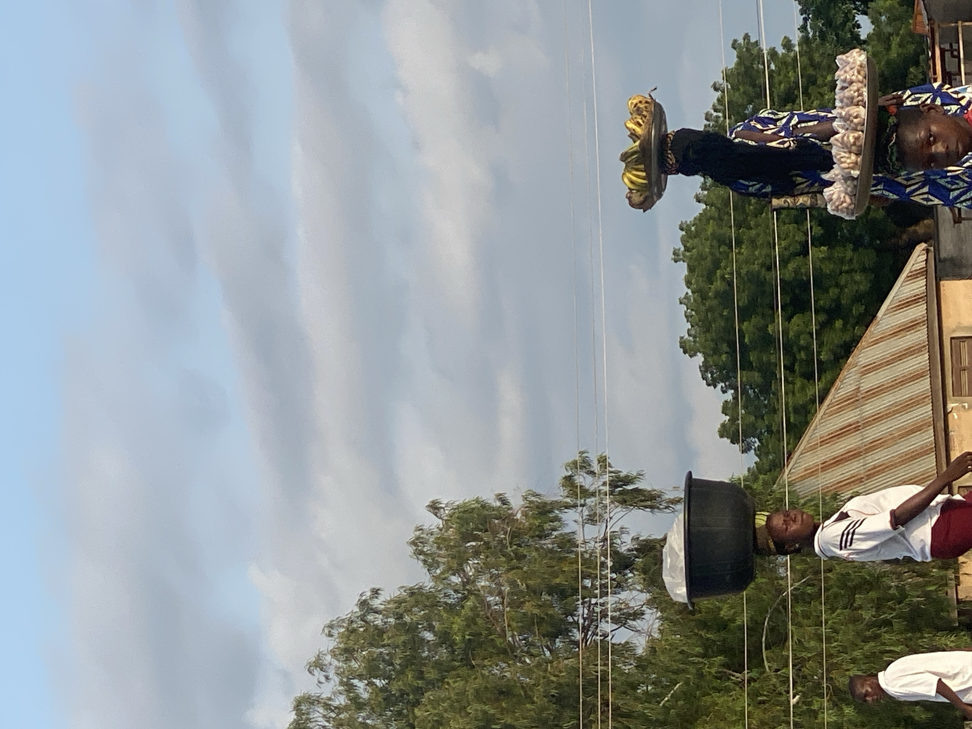 In Jalingo, Nigeria, a person is depicted balancing a large black tub on their head, filled with what appears to be plastic sheeting. They are wearing a white shirt with black stripes on the sleeves and dark maroon shorts. The person is positioned outdoors, with a background of lush green trees and a partially visible building with a corrugated metal roof. Another person, also wearing a white shirt, is partially visible in the lower right of the frame, seemingly walking or standing behind the first individual. To the left of the main subject, another person is seen balancing a tray on their head, laden with bunches of bananas and what looks like a bundle of garlic or onions wrapped in plastic. This individual is wearing a vibrant blue and white patterned fabric. The scene suggests a marketplace or street setting, possibly during the daytime given the lighting. The sky is a pale blue with scattered clouds. There is no visible text in the image.