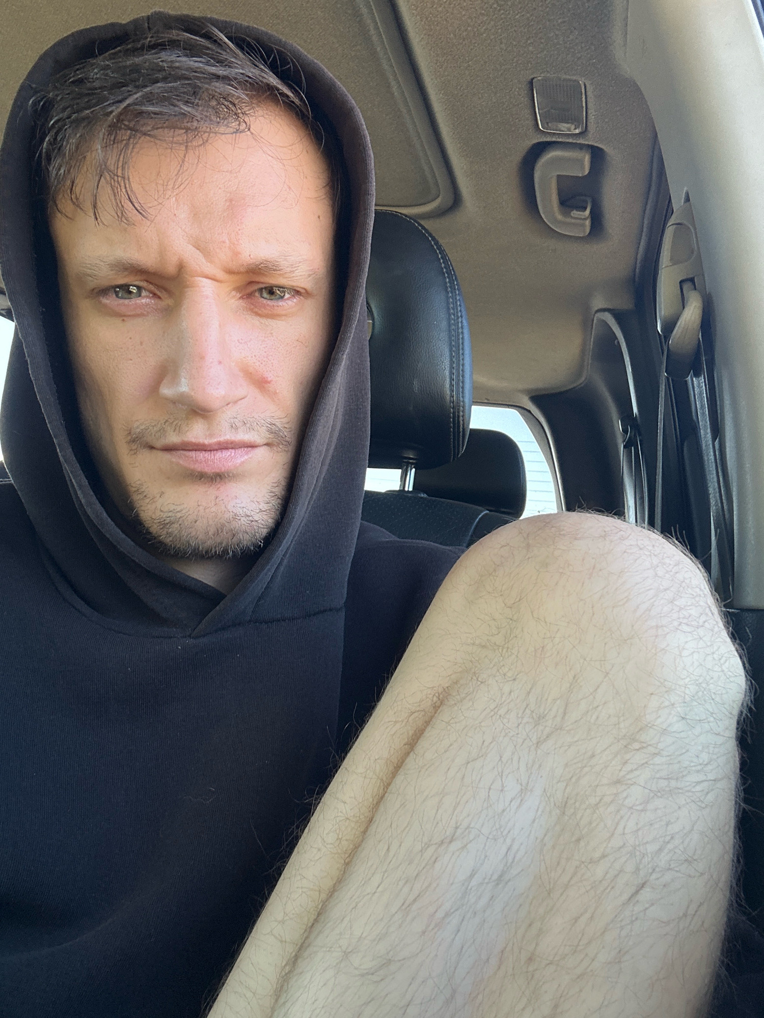 The image shows a close-up, selfie-style shot of a man with light skin, dark hair, and a beard stubble, wearing a black hooded sweatshirt. He is seated inside a vehicle, with part of his leg visible in the foreground, showing light hair. The interior of the vehicle appears to be a standard car interior, with a headrest, seatbelt anchor, and part of the car's roof lining visible. The man is looking directly at the camera with a serious expression. The lighting suggests it is daytime. The scene is consistent with someone taking a photo from the driver's seat of a car. There is no indication of the specific city of Makiv, Ukraine, in the image itself.