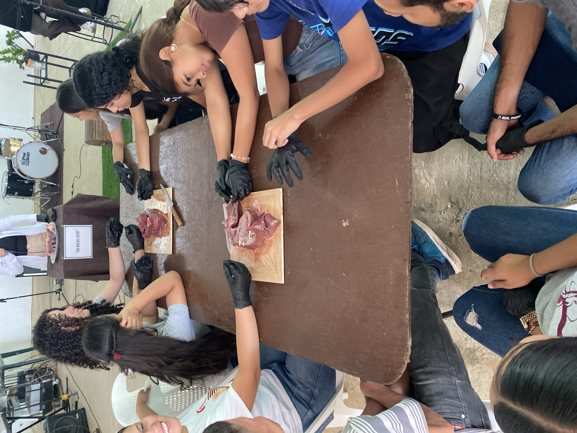 In Tinaquillo, Venezuela, a group of people wearing black gloves stand around a brown wooden table. At least 10 individuals are visible, including males and females. The participants are performing a dissection, evident by the presence of raw organs placed on wooden boards. A sign reading "Un Nuevo Latir" is present. Additional details in the environment include a drum set in the background and a table displaying more organ meat.