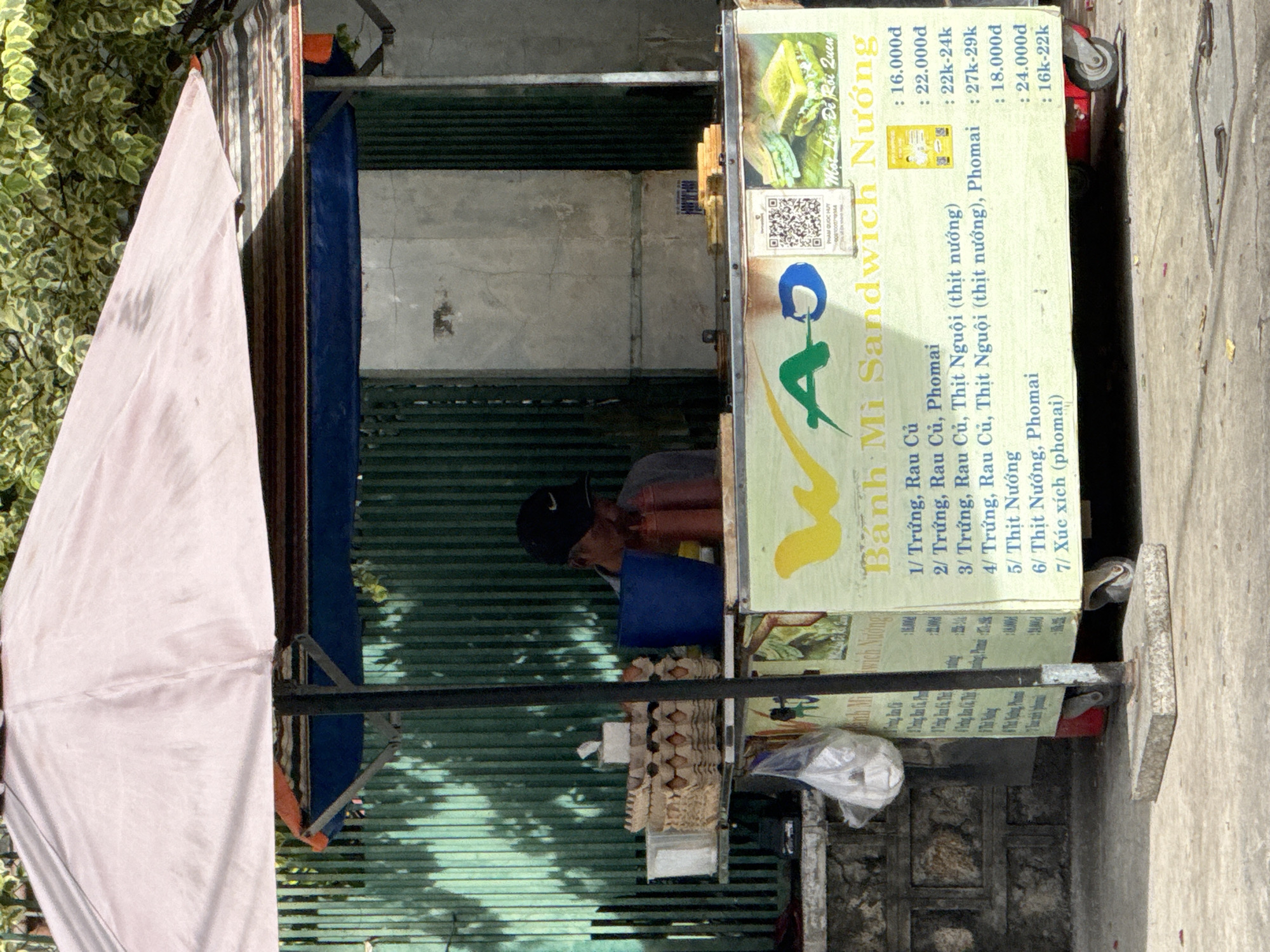 A street vendor is selling banh mi sandwiches from a cart under a pink umbrella. The cart is located in front of a green corrugated metal fence and a concrete wall. A man wearing a black baseball cap is standing behind the cart, out of focus. The cart has a yellow sign with Vietnamese text and prices for different sandwich options, as well as images of sandwiches. Several egg cartons and a blue bucket are visible on a shelf in the cart. The ground in front of the cart is concrete, with some debris scattered on it. The scene appears to be a typical street food setup in Vietnam.