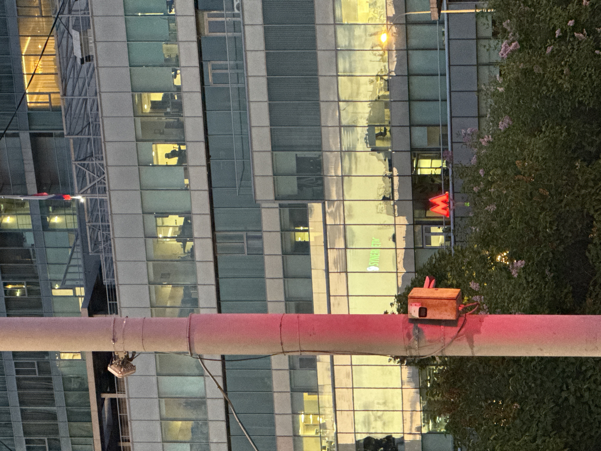 The image captures an urban scene, likely in Moscow, Russia, identifiable by the presence of the Moscow Metro "M" logo in red, situated against a multi-story building with a modern glass facade. The building reflects light and the surrounding environment. In the foreground, a horizontal, light-colored cylindrical object, possibly a pipe or beam, runs across the image. Attached to this object is a wooden box with an unknown function. To the right of the box, foliage from a tree or bush is visible. The scene is partially obscured by the foreground elements, but the architectural details and the presence of the Metro logo indicate a developed urban setting. There are no people visible in the image. The time of day appears to be dusk or evening, suggested by the lighting conditions.