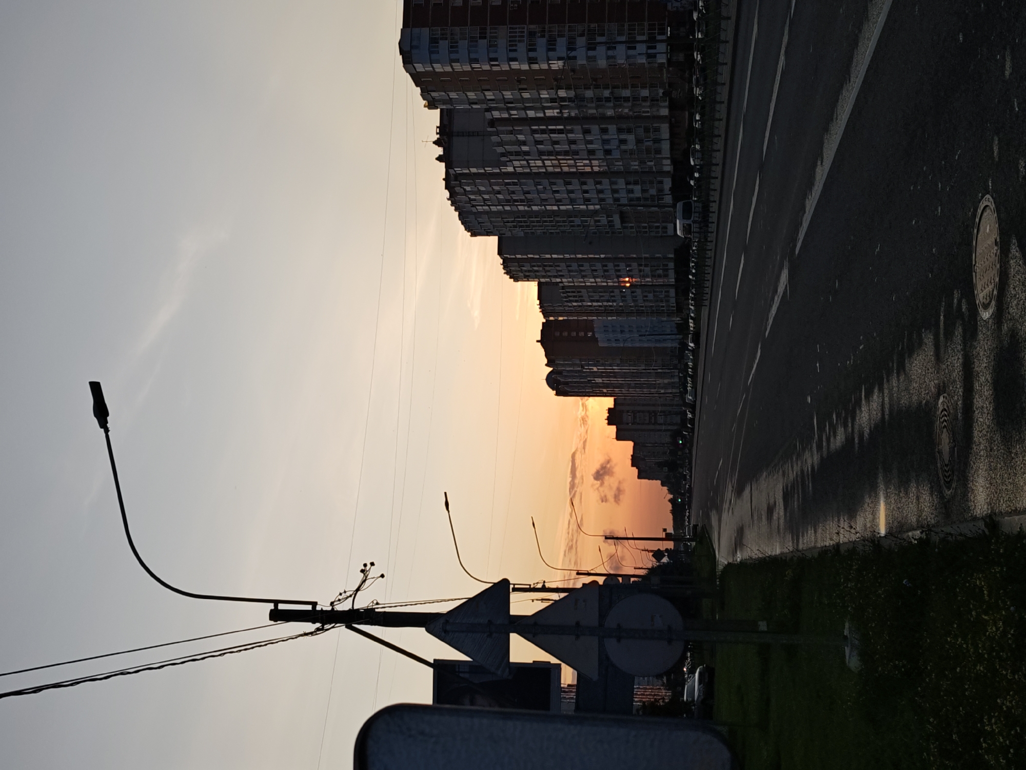 The image captures a dusk scene along a wide urban road in Saint Petersburg, Russia. The sky displays a gradient of soft yellow and orange hues near the horizon, indicative of sunset, transitioning to a pale blue overhead. A thin, wispy cloud streaks across the sky.
On the right side of the road, a row of multi-story residential buildings, typical of Saint Petersburg's urban landscape, stretches into the distance. These buildings are characterized by their uniform facades and numerous windows, most of which are dark. A few streetlights are visible along the road, silhouetted against the sky. The road itself is paved and appears wet in places, reflecting the fading light. Lane markings are clearly visible on the asphalt.
On the left side of the road, a grassy verge is present, with a few low-lying plants. A prominent traffic light pole with multiple wires and attached streetlights stands near the foreground. Several traffic signs, including triangular warning signs, are mounted on poles next to the streetlights. A large, indistinct object, possibly a vehicle or a temporary structure, partially occupies the far left of the frame. Cars are parked or moving slowly along the road in the distance. The overall impression is of a typical city thoroughfare during twilight.