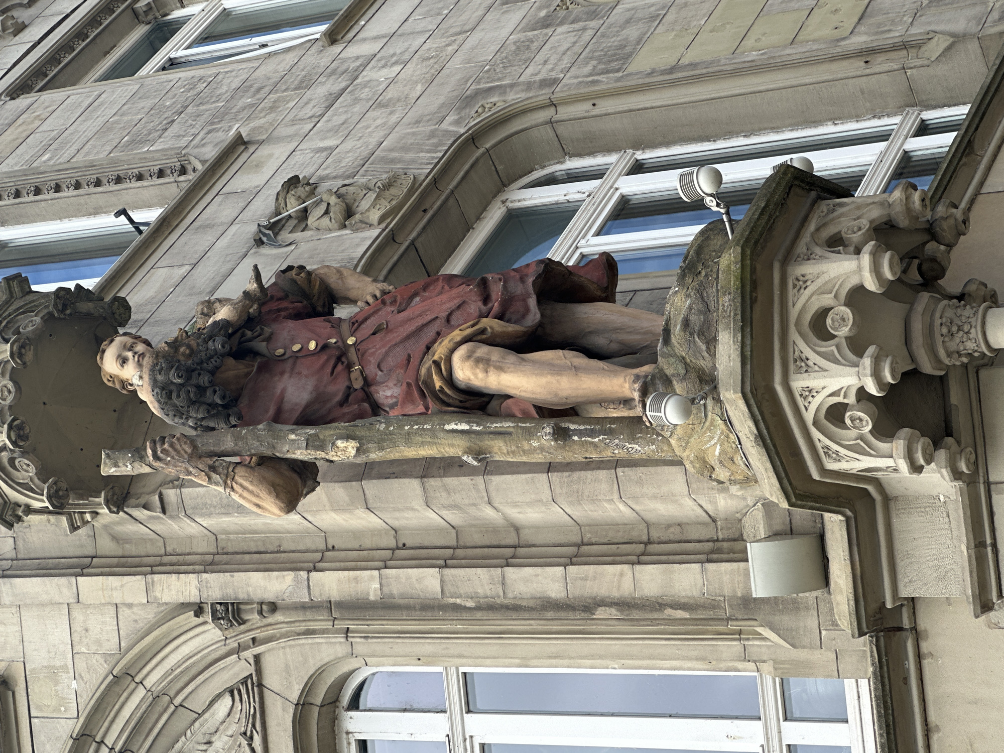 The image depicts a large, multi-colored statue affixed to the facade of a light grey stone building under an overcast sky. The primary statue, positioned on an ornate stone corbel, features two figures: a towering adult male in a reddish-brown tunic and a lighter-skinned child perched on his shoulders. The adult male, facing slightly left, grasps a thick, branch-like staff with both hands, its lower end obscured. His tunic is detailed with gold buttons and a belt, and his lower legs are bare. The child, looking directly forward, has light hair and skin.
The building facade is constructed from large, rectangular stone blocks, showing signs of weathering and vertical staining in some areas. Several arched windows with white frames are visible, reflecting a pale blue and white sky. Architectural details include decorative cornices, moldings, and smaller carved figures. To the right of the main statue, a smaller monochrome carving of a figure holding a tool (possibly a pickaxe or hoe) is present, with a banner below it appearing to bear the German word "BAUER" (farmer). Further right and higher, another decorative frieze of repeating rosette patterns runs horizontally along the building.
Modern fixtures are integrated into the facade around the statue. Two white, dome-shaped sensors or lights are mounted on silver metal arms; one is located just below and to the right of the statue's feet, and another is further to the right, lower, and closer to a window. A rectangular grey utility box is affixed to the stone wall below the statue's base, to the left. A black metal bracket is visible above and to the right of the statue's head.
The location is Unknown, Unknown.