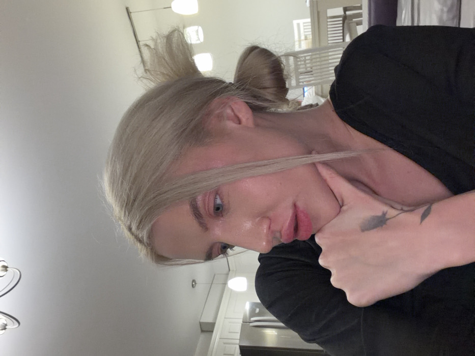 This image depicts a single individual, identified as a female, within an indoor residential setting in Budva, Montenegro. The subject has blonde hair pulled back into a bun with some strands framing the face, blue eyes directed downwards and to the left, and full lips enhanced with a pink hue. She wears a black v-neck shirt. Her right hand, from the viewer's perspective, supports her chin; a dark floral tattoo is visible on her right wrist/forearm. The background features a modern kitchen and dining area. Visible elements include white ceiling, a chandelier, a stainless steel double-door refrigerator on the left, white kitchen cabinetry, and a white dining table with matching slatted-back chairs on the right. A portion of a dark-colored cushioned surface is present in the lower right foreground.