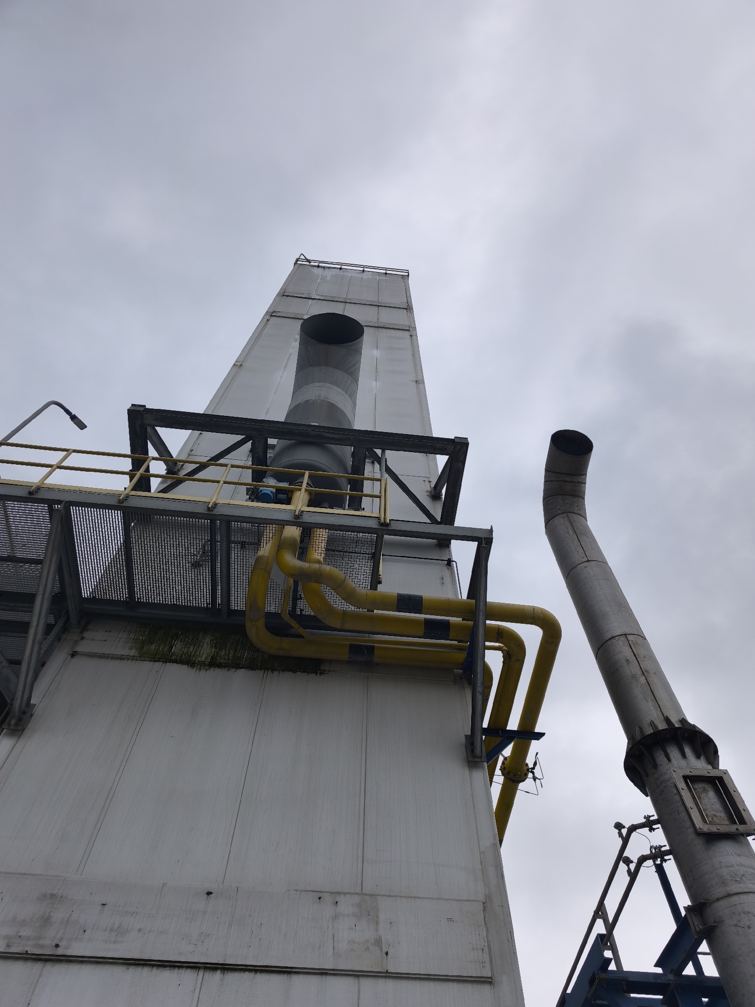 The image presents a low-angle view of large industrial structures against an overcast, grey sky. The central focus is a tall, rectangular, light-colored building or tower, appearing to be made of paneling. Attached to this structure, at about a third of its height from the bottom of the frame, is a multi-level platform system with metal grating and yellow railings. A metal staircase is visible on the left side of the platform.
Numerous yellow pipes run across and along the platform, bending and connecting, suggesting a network for transporting fluids or gases. A large, grey, vertical exhaust stack emerges from the top of the main tower, with its opening facing upwards. Below the platform, on the lower part of the main structure, there is visible dark green and black grime or algae accumulation.
To the right of the main tower, another prominent, large, grey metallic pipe or stack rises diagonally upwards. This pipe has a distinct bend at its top section, curving towards the upper right, and features an access panel or inspection plate, along with various smaller pipes and fittings attached to its lower visible section. A small blue component, possibly a valve or sensor, is visible on the platform near the central stack. The sky is uniformly grey and cloudy, indicating an overcast day. No human activity or visible text is present in the scene. The overall impression is one of a heavy industrial facility.