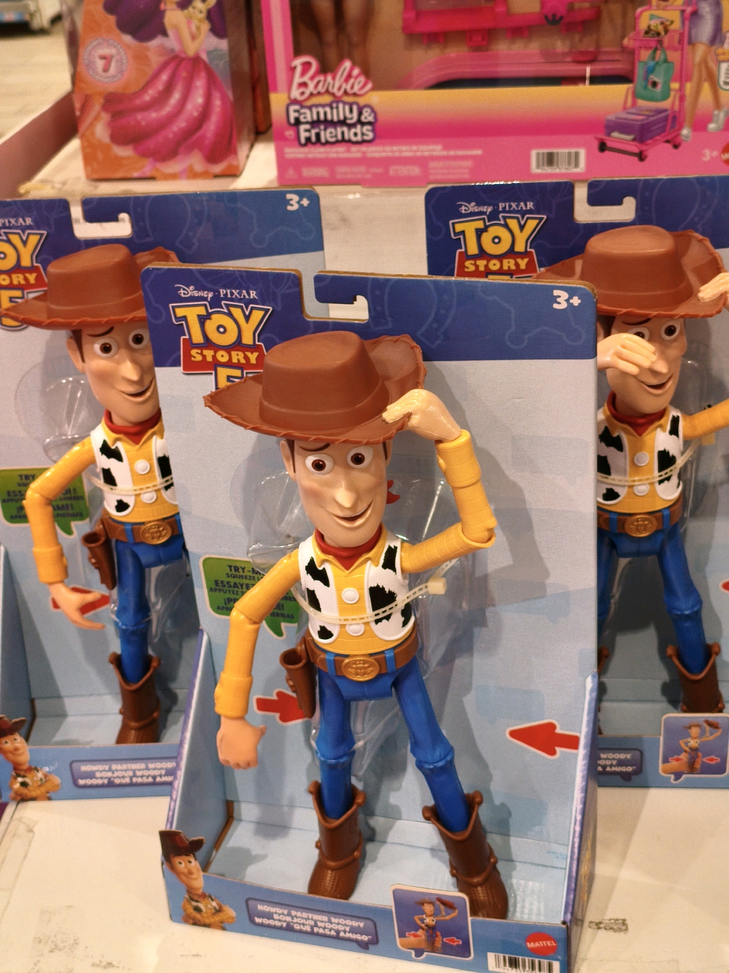 In Babakan Madang, Indonesia, a retail display features multiple "Toy Story" Woody dolls in their packaging. Three full-sized Woody dolls are visible, each wearing his signature cowboy attire: a yellow long-sleeved shirt, a white vest with black spots, blue pants, and brown cowboy boots and hat. They are positioned in front of blue cardboard packaging adorned with the "Toy Story" logo. Two of the dolls are closer to the foreground, with one slightly behind and to the right of the other. A smaller Woody figure in packaging is visible on the bottom left of the image, and another is partially visible on the bottom right. In the background, out of focus, are toy boxes, including one with a Barbie advertisement and another featuring a cartoon princess in a pink dress. The scene appears to be within a store setting.