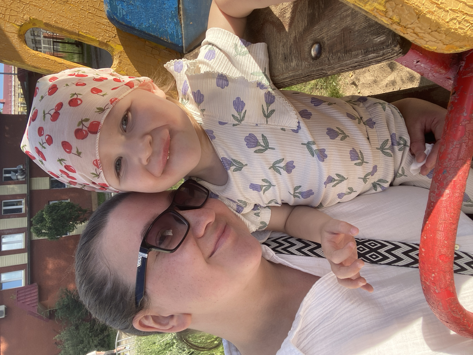 This media file shows a close-up, sideways selfie of a woman and a young child. The woman, wearing glasses and a white textured shirt, is in the foreground. Her expression is relaxed, and she appears to be smiling faintly. Behind her, a young child is nestled close, wearing a white bandana with a cherry print and a light-colored outfit patterned with purple flowers. The child has a wide, joyful smile.
They appear to be on a playground, with parts of a colorful structure visible, including yellow, blue, and red painted surfaces and wooden planks. The background shows greenery and a brick building with multiple windows, suggesting an outdoor urban or suburban setting. The lighting indicates it is daytime, likely sunny, casting shadows and highlighting the textures of the clothing and playground equipment. The overall mood is one of happiness and connection between the two individuals. No visible text is present in the image.