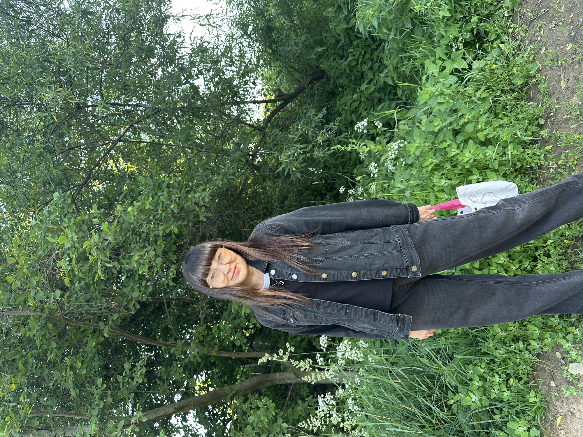 A young woman with dark hair and bangs, wearing glasses, a black t-shirt, a black denim jacket, and black jeans, poses in a natural outdoor setting. She is holding a pink umbrella and a white bag with illustrations on it. The background consists of lush green trees and foliage, with some white wildflowers scattered in the grass. The lighting suggests it is daytime, possibly late afternoon or early evening, with soft natural light. The overall mood is relaxed and casual. The image appears to be taken in Chaikovsky, Russia, given the location context. There is no visible text in the image.
