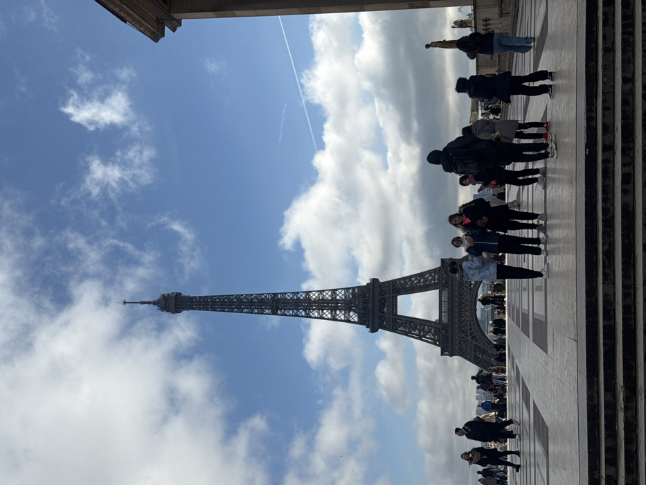The image displays a daytime view of the Eiffel Tower in Paris, France, observed from the Esplanade du Trocadéro, part of the Palais de Chaillot complex. The iconic iron lattice tower dominates the left-center background, its full structure visible against a blue sky with scattered white cumulus clouds. A faint linear aircraft contrail extends horizontally across the upper right sky.
In the foreground, a wide, multi-tiered paved platform with dark, broad steps transitions into a patterned, lighter-colored esplanade. Approximately twenty individuals are present throughout the scene. In the midground, a cluster of about ten people, primarily young adults, stands on the raised esplanade. Some are holding mobile phones, while others appear to be conversing or observing. Their attire includes dark jackets, long pants, and sneakers, suggesting cool weather. Further into the background, numerous smaller figures are dispersed across the plaza leading towards the base of the Eiffel Tower. On the far right, a segment of a classical stone building with a prominent cornice is visible, along with a dark, standing statue on a pedestal. Actions observed include standing, walking, and passive observation of the surroundings.