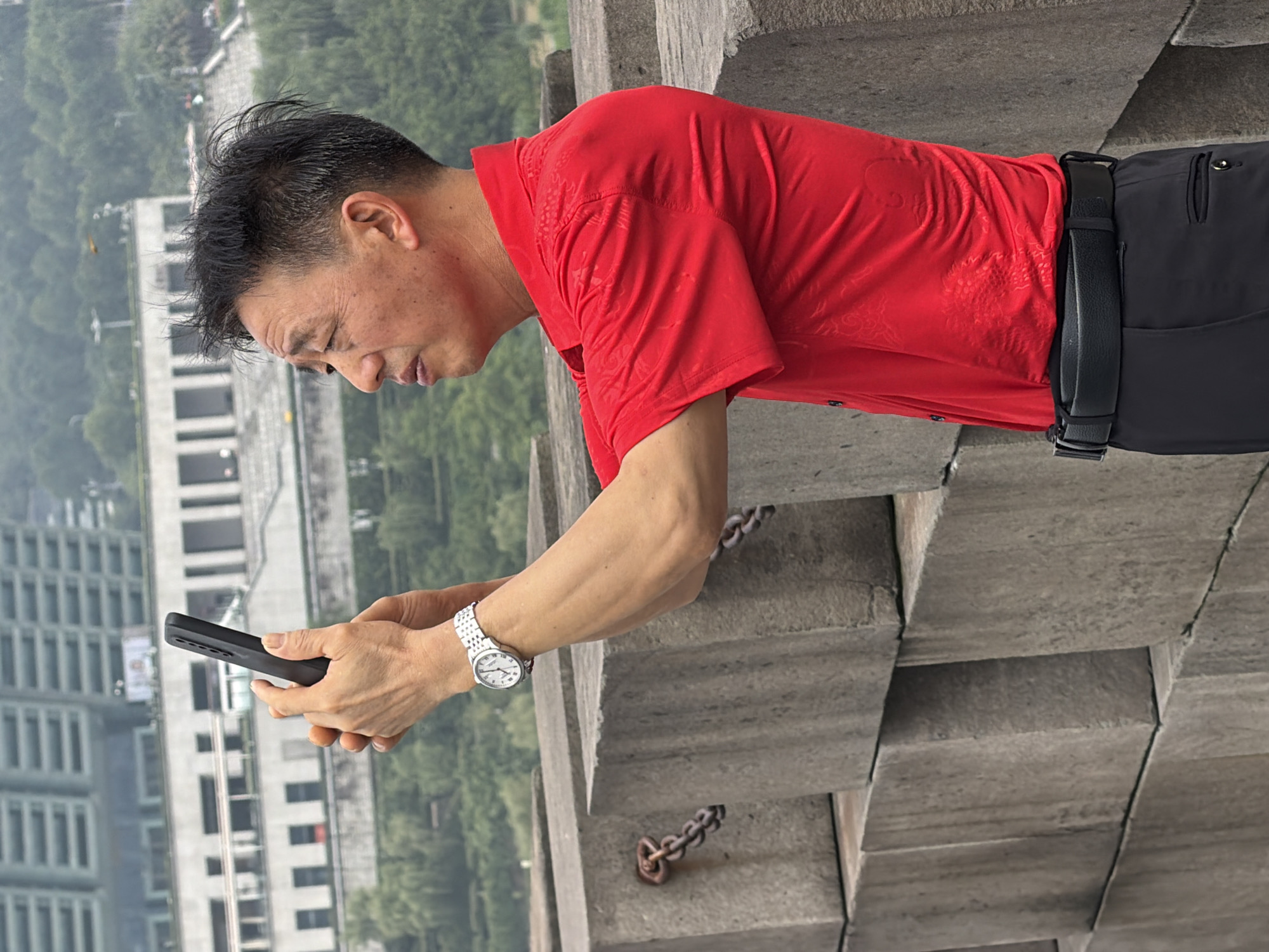 An East Asian man with short, dark, messy hair is standing on a raised platform of large concrete blocks, holding a black smartphone in his right hand, which is extended forward as if taking a selfie. He is wearing a bright red short-sleeved collared shirt with a subtle embossed pattern, a black belt, and black pants. A silver-banded wristwatch is visible on his left wrist. The background shows a blurred cityscape with modern buildings and lush green trees. The concrete blocks in the foreground have metal chain links attached to some of them. The scene appears to be outdoors in an urban environment.