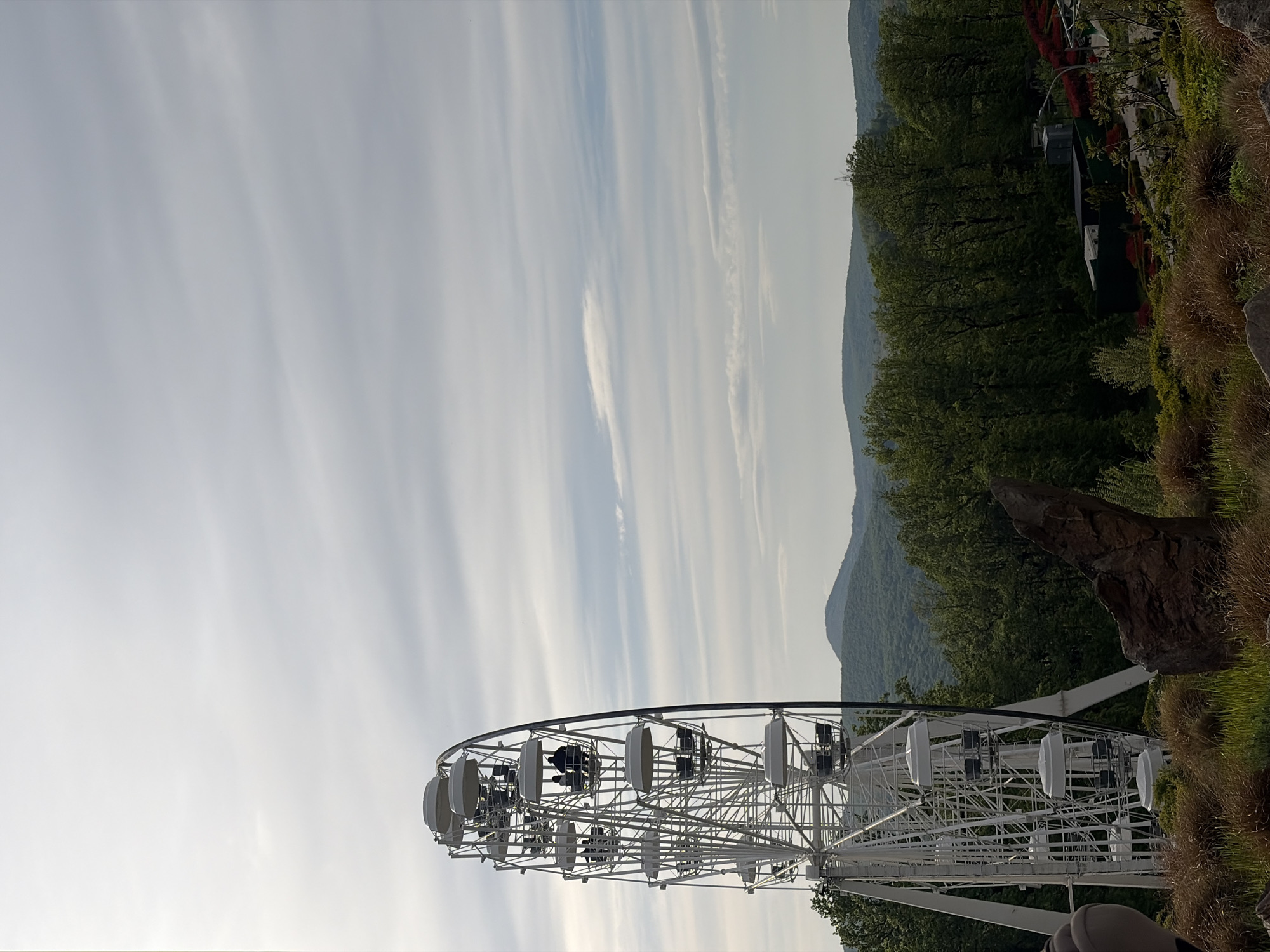 The image depicts a Ferris wheel in Goryachiy Klyuch, Russia. The white Ferris wheel is positioned against a backdrop of a partly cloudy sky, rolling hills, and dense green vegetation. Passengers are seated in the gondolas. The foreground includes some ground cover and a large rock formation.