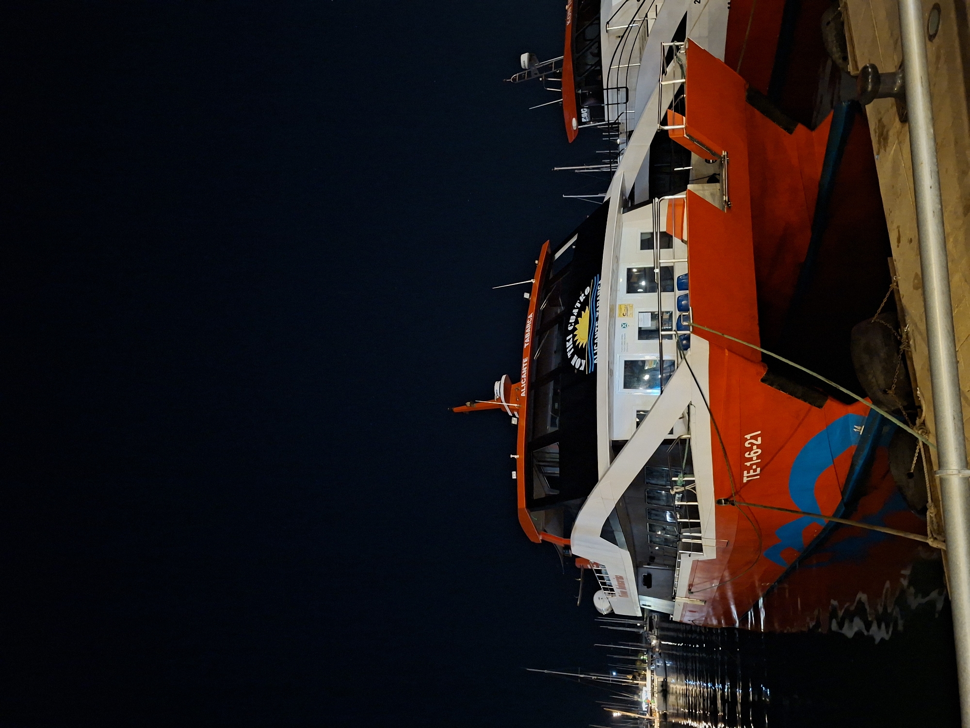 This image shows two modern passenger catamarans docked at night in the port of Alicante, Spain. The foreground is dominated by the closest catamaran, painted bright red with a white superstructure and blue wave-like graphics on its hull. The vessel has the registration number "TE-1-6-21" clearly visible on its side. Above the main deck, the superstructure is white with large windows. Text on the windshield reads "ALICANTE TABARCA" and "KON TIKI CUATRO," indicating it is a ferry service to the island of Tabarca. Blue and orange seats are visible through the windows.
To the right of the foreground vessel, a second, similar catamaran is partially visible. It also has an orange trim around its upper deck and white hull. The text "KON TIKI" is discernible on its side.
The background consists of the dark night sky and the dimly lit port area. Several masts of sailboats and other vessels are visible in the distance to the left, suggesting a marina. The water in the foreground reflects the lights from the boats and the port, creating shimmering patterns. The scene is calm, with no people visible. The overall impression is one of a quiet night in a bustling port city.