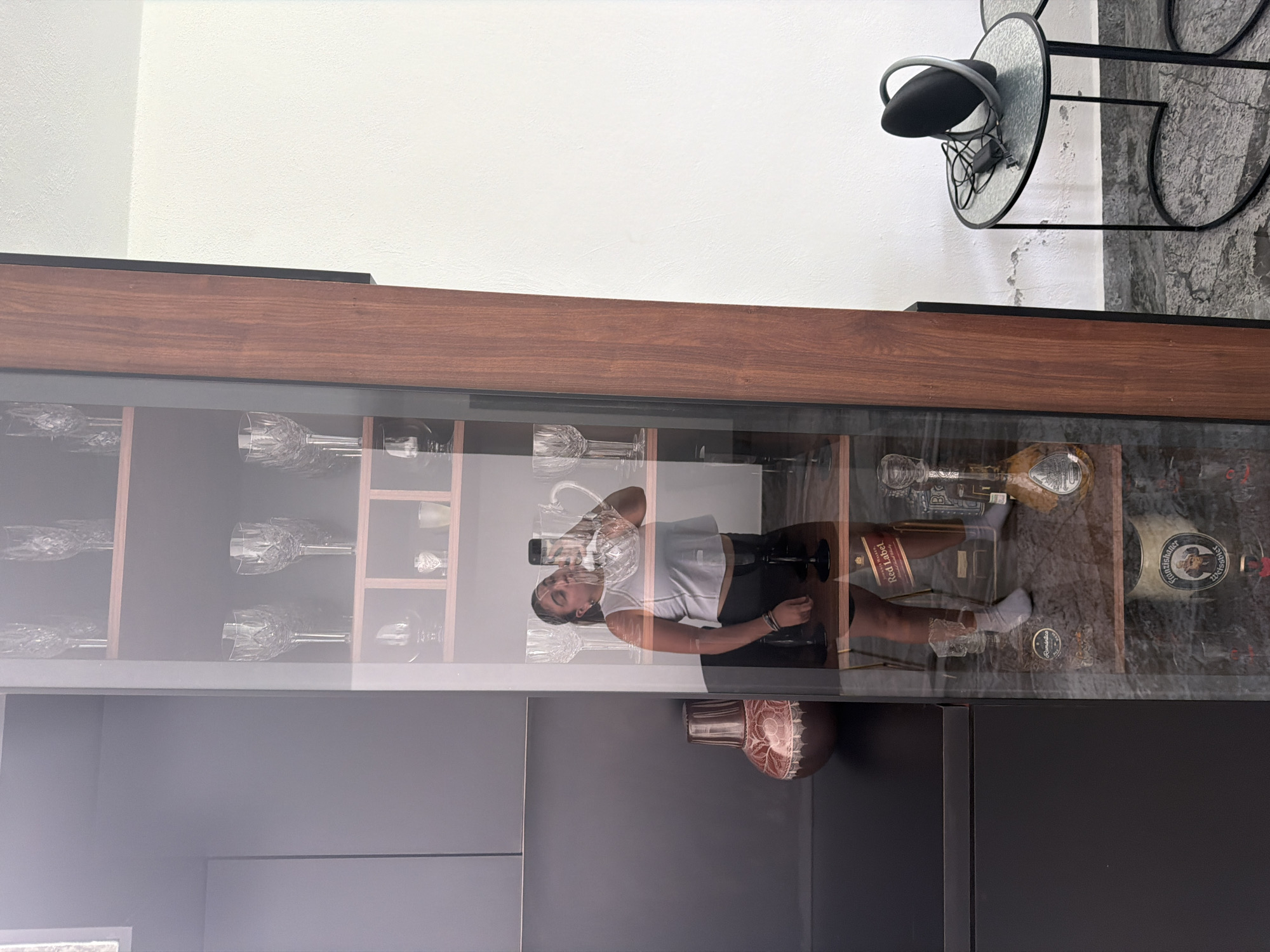 The scene depicts a modern kitchen cabinet with glass doors, likely in a residence in Puebla, Mexico. The cabinet is a combination of dark gray and wood finish. The cabinet’s glass doors reflect an image of a young woman taking a photo of herself with her phone. The woman has dark hair, wearing a white top and dark shorts. She is standing in front of the cabinet, capturing her reflection. Inside the cabinet are various wine glasses, liquor bottles (such as “Johnnie Walker Red Label” and “Paulaner”) and other items. Some of the items are reflected in the glass, obscuring the details. The environment appears to be indoors, with a white wall visible above the cabinet and a circular black table and chair setup partially seen at the top right.