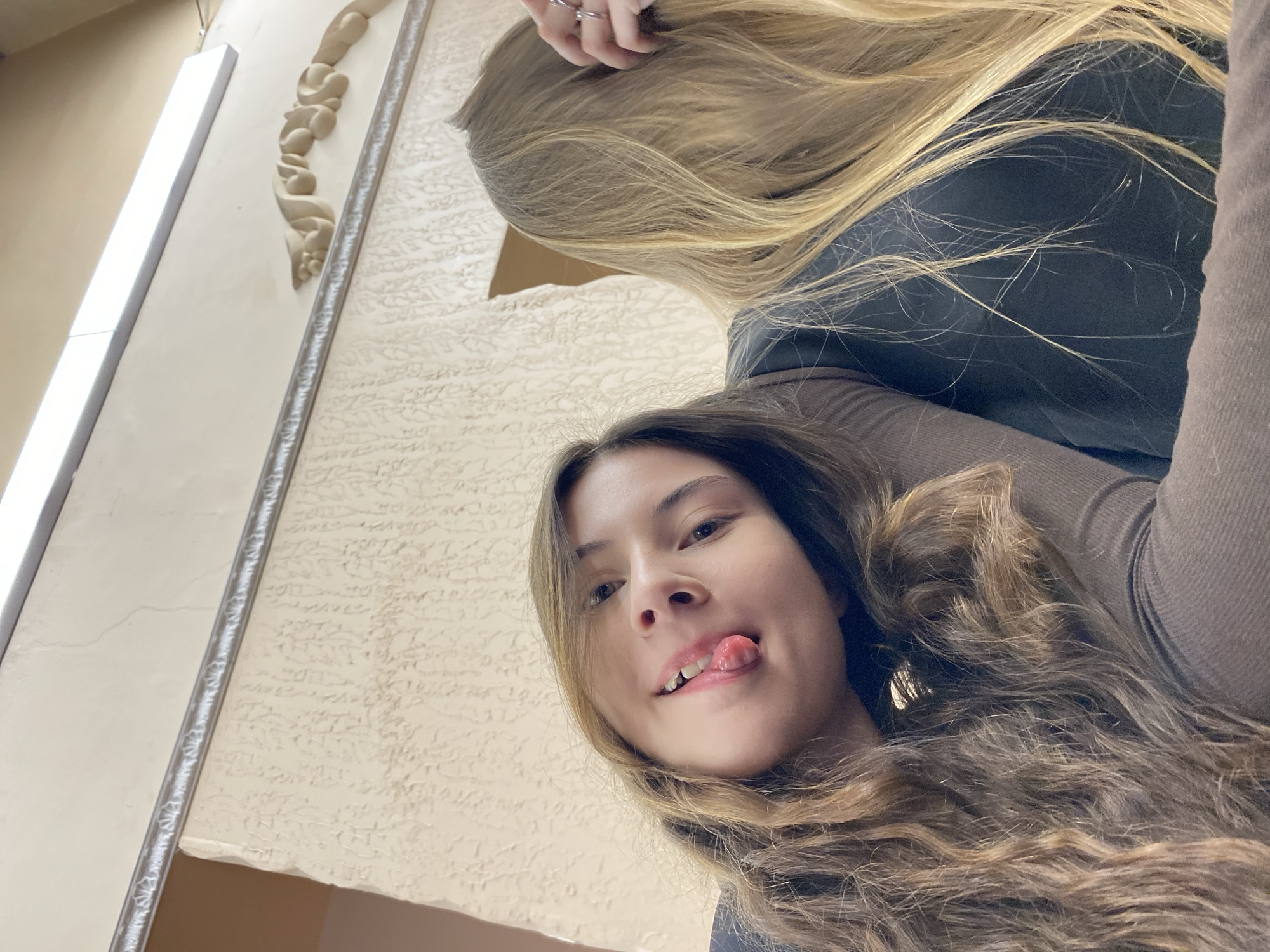 Two women are in the image, taken from a low angle. One woman has long, wavy, light brown hair with blonde highlights, and her tongue is sticking out. The other woman has long, straight, blonde hair. They are both wearing dark-colored shirts. The background is a light-colored wall with textured plaster. A decorative molding is on the wall, and a fluorescent light fixture is visible. There is no explicit information about the location being in Votkinsk, Russia.