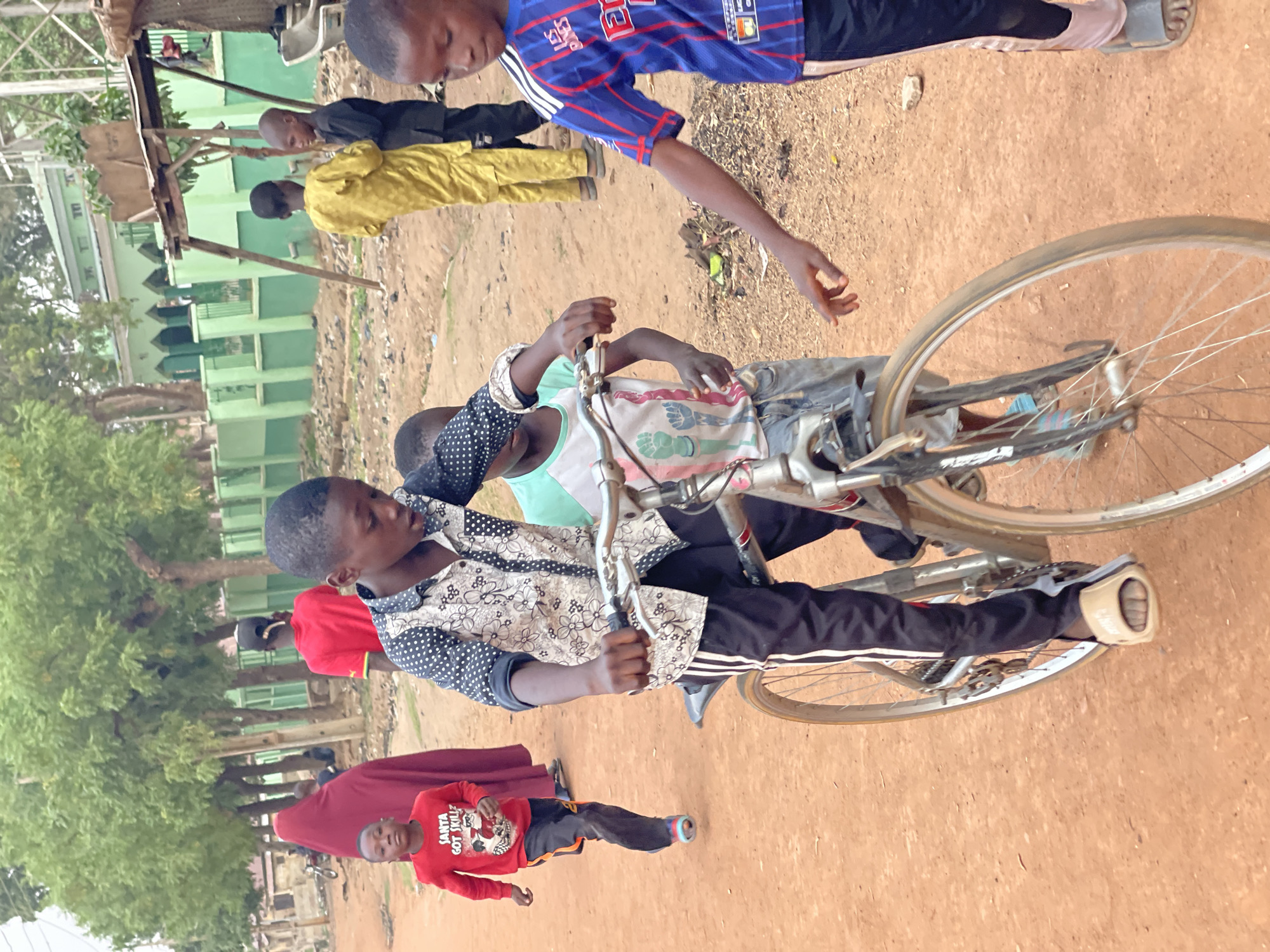 In the city of Zaria, Nigeria, a group of children are seen. One child is riding a bicycle. Another child stands nearby with his hand reaching out towards the bicycle. Other children stand and walk in the background. The environment includes a dirt or sandy ground, trees, and a building. The building appears to be green. One child wears a red shirt with the text "Santa Got Skillz". Another child wears a blue and red jersey. The boy on the bicycle wears a patterned shirt and black pants with a white stripe down the side.