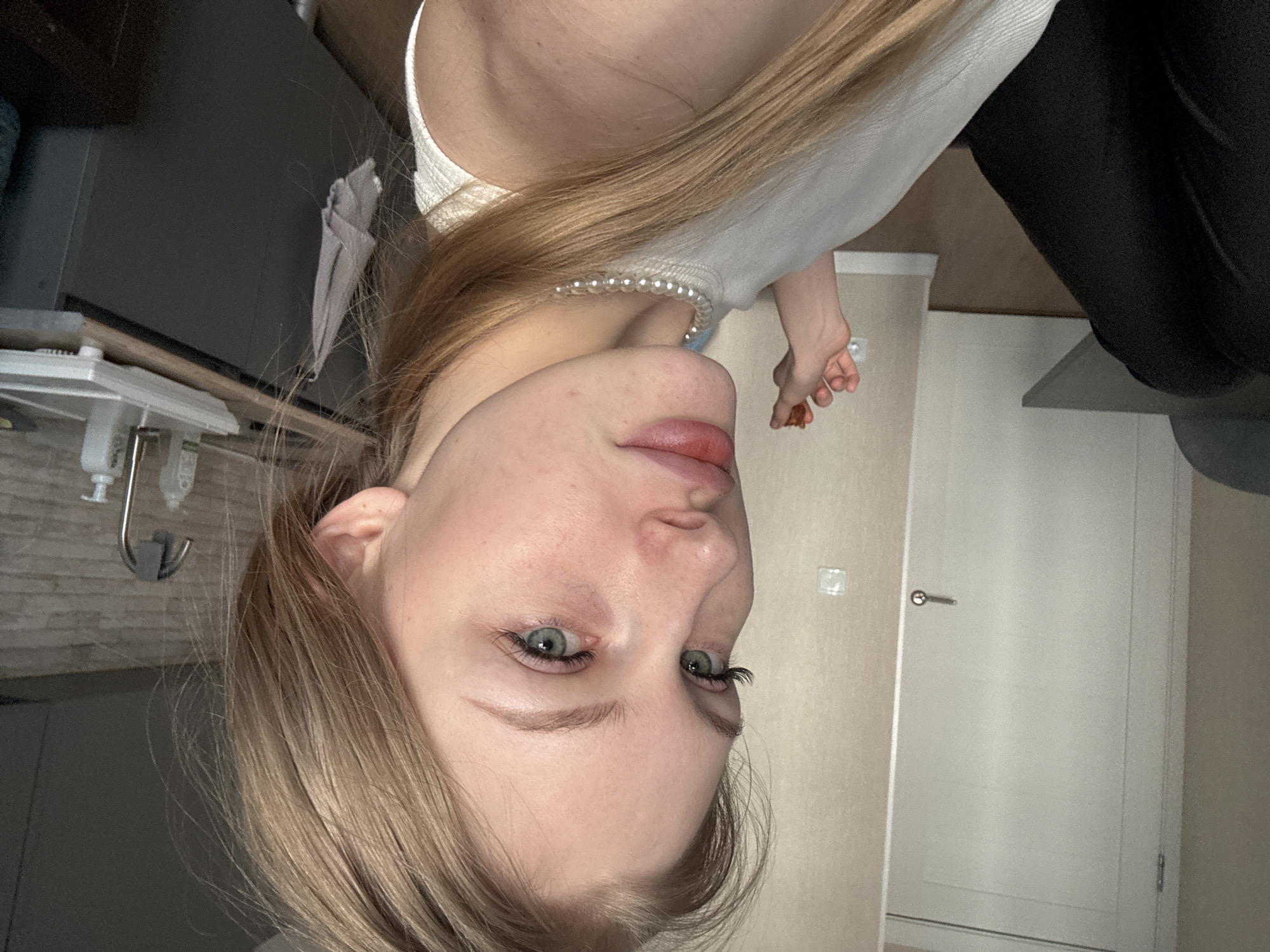 A young woman with long, light brown hair and blue-green eyes looks directly at the viewer, her lips slightly pursed. She is wearing a white top and a simple pearl necklace. Her right hand is visible, holding a small, reddish-brown, irregularly shaped object, possibly a piece of food. The setting appears to be indoors, likely a home. To the woman's left, a light-colored interior door with a silver handle and a wall-mounted light switch or electrical outlet are visible against a beige wall. To her right, partially visible elements suggest a kitchen area, including what might be a sink faucet and a soap dispenser. A dark, indistinct object, possibly a chair or furniture, is in the upper right background. The general lighting suggests it is daytime. The image, originally rotated 90 degrees counter-clockwise, depicts a casual indoor moment in Perm, Russia. There is no visible text.