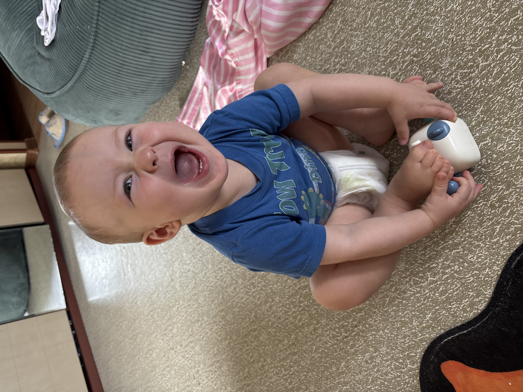 An infant sits cross-legged on a speckled carpet in what appears to be a home environment. The baby wears a blue t-shirt with a graphic print and a diaper. The infant's mouth is wide open in a scream or laugh, and their eyes are squeezed shut. One foot is positioned in front of them, with small hands reaching out to touch a white and blue toy. A pink and white striped blanket is visible to the right of the infant. In the background, a large, dark green corduroy beanbag chair is present, along with what looks like part of a cabinet with mirrored doors. The overall lighting suggests it is daytime, with natural light possibly coming from a window. There is no discernible indication of the city of Naberezhnye Chelny, Russia.