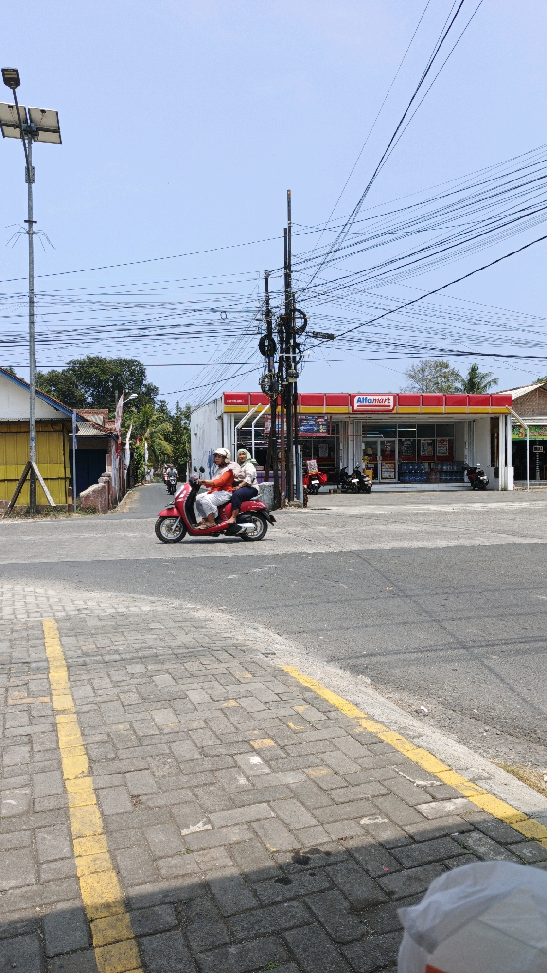 In the city of Cibenda, Indonesia, a street scene unfolds under a clear blue sky. Two individuals are riding on a red scooter, moving from left to right across the frame. The rider in front wears an orange long-sleeved shirt and white pants, with a white helmet. The passenger behind them is wearing a light-colored long-sleeved shirt and a light green headscarf, also with a white helmet. They are positioned on a paved road with distinct yellow markings.
To the right of the scooter is an Alfamart convenience store, a brightly lit building with a red and yellow facade. Several motorcycles are parked in front of the store. In the background, more electrical poles with a complex network of wires crisscross the sky, a common sight in urban Indonesian landscapes. On the left side of the image, there are buildings, including one with a yellow wall, and trees are visible further in the distance. The foreground shows a paved sidewalk with a yellow line running along its edge. The overall scene depicts a typical day in an Indonesian town.