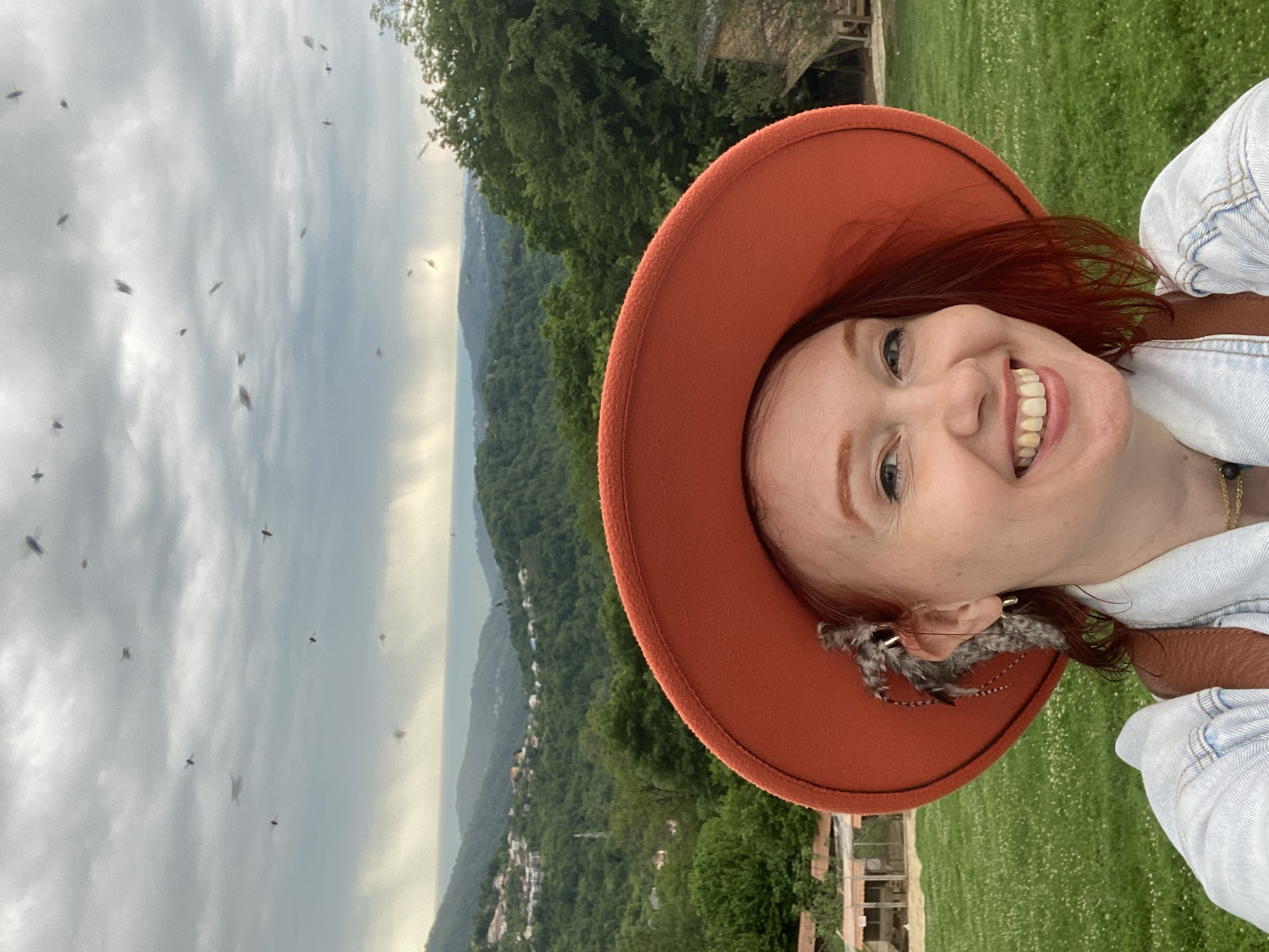 A woman with red hair, wearing a tan hat with a feather decoration, smiles in a selfie. The background shows a cloudy sky with many small flying insects, a hillside covered in trees, and buildings in the distance. The location is unspecified, but based on the prompt it is said to be the city of Прогресс, Russia.