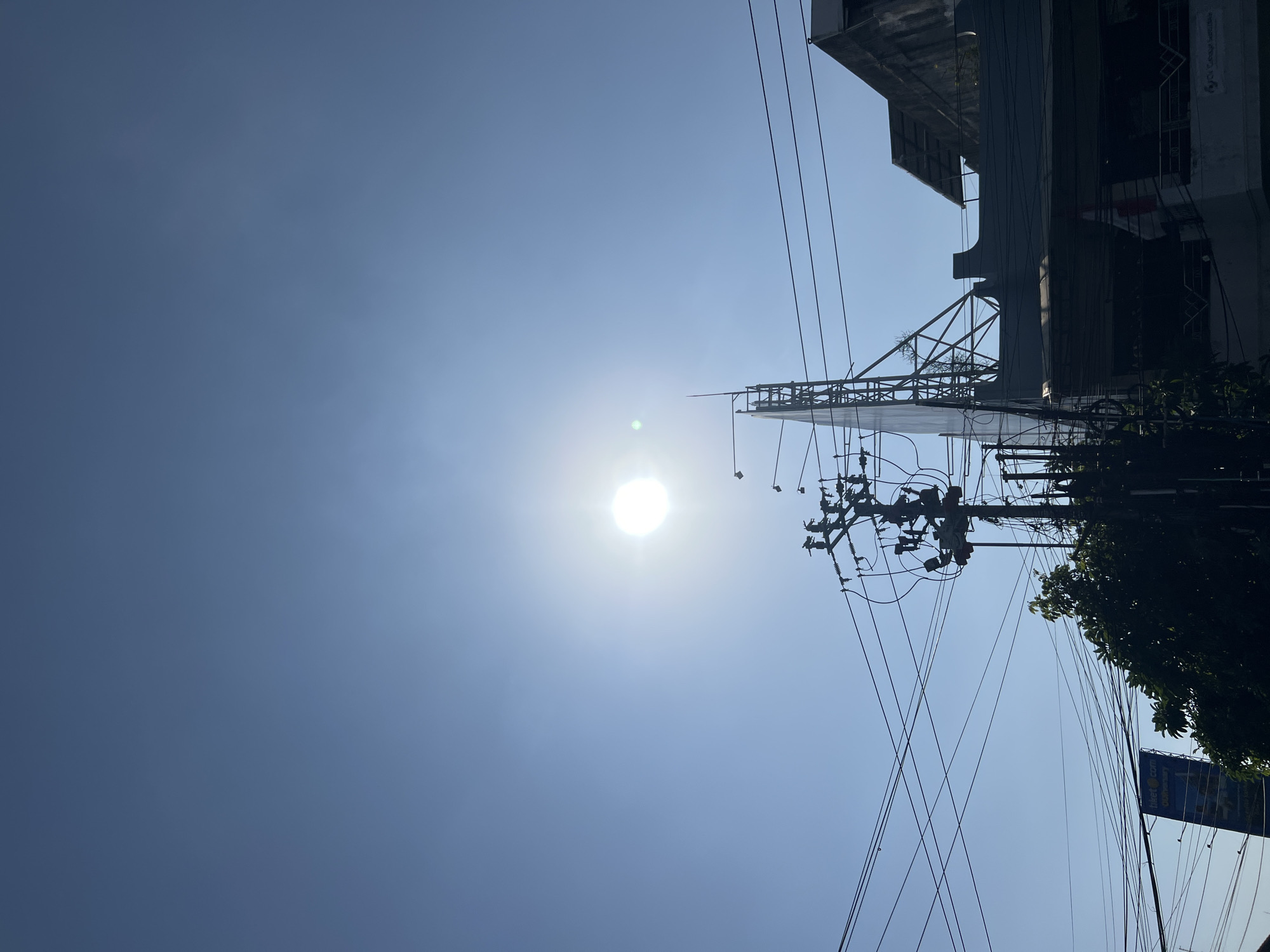 A wide shot captures a clear, bright blue sky over Denpasar Barat, Indonesia, on a sunny day. The sun is prominently positioned in the upper center of the frame, its intense light creating lens flare. A utility pole with numerous electrical wires is visible in the middle ground, silhouetted against the sky. To the right of the pole, a large billboard structure with a white face and metal scaffolding stands tall. The upper floors of a building with a dark roof and balconies are visible on the far right. Dense green foliage from a tree peeks out from behind the utility pole and billboard on the left side. A blue billboard with white text advertising "tiket.com" is partially visible in the lower left corner. The overall scene depicts an urban streetscape under a bright, cloudless sky.