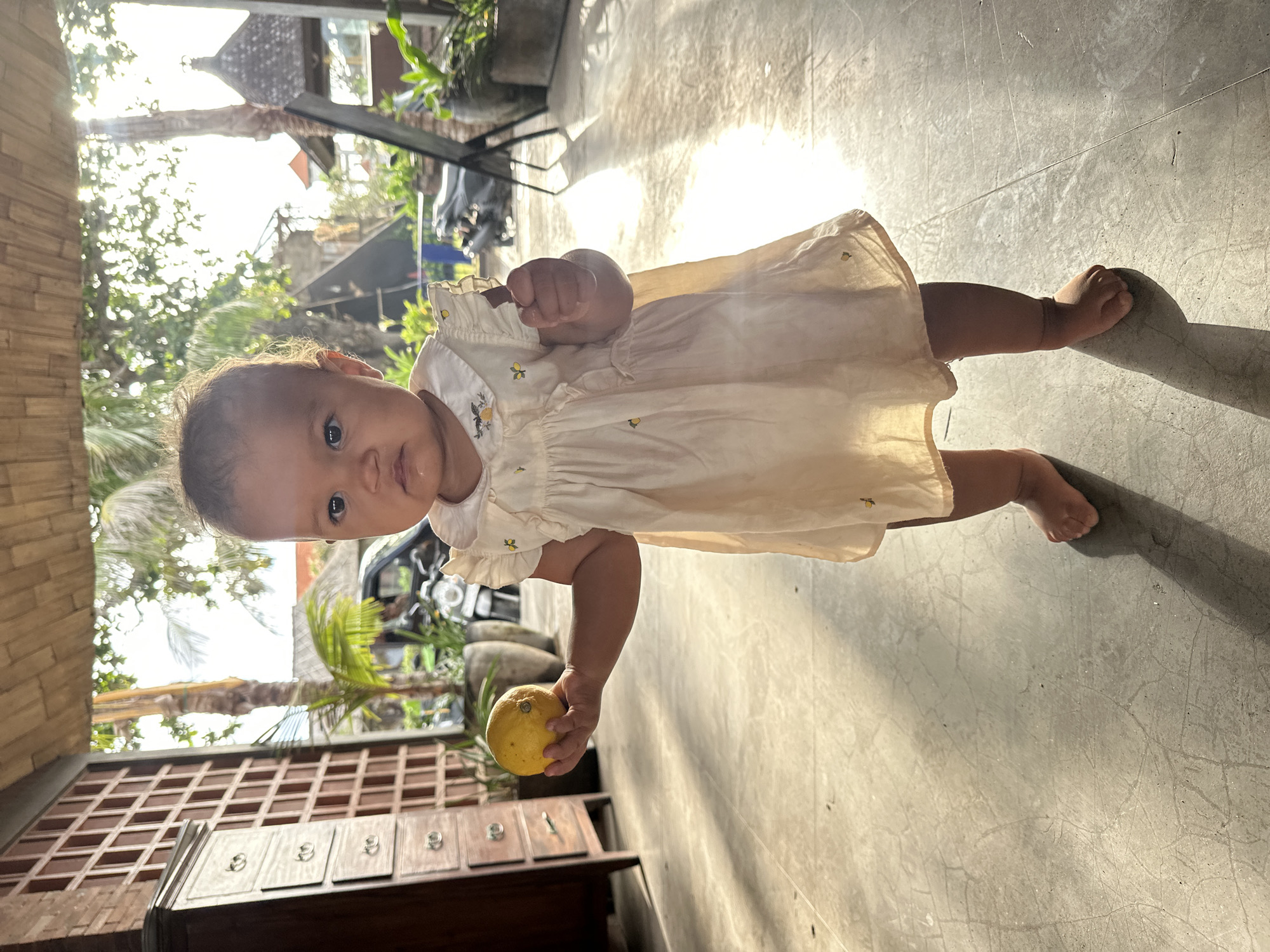 A young girl stands on a concrete floor, holding a yellow fruit in her left hand. She is wearing a light-colored dress with ruffles on the sleeves and a collar decorated with small embroidered lemons and birds. Sunlight streams from the right, casting shadows on the floor and illuminating the girl. In the background, a wooden cabinet with drawers is visible to the left, and beyond that, a doorway opens to an outdoor area with palm trees and a motorcycle. The setting appears to be an open-air structure, possibly a cafe or guesthouse, in North Kuta, Indonesia, characterized by its tropical vegetation and rustic architecture.