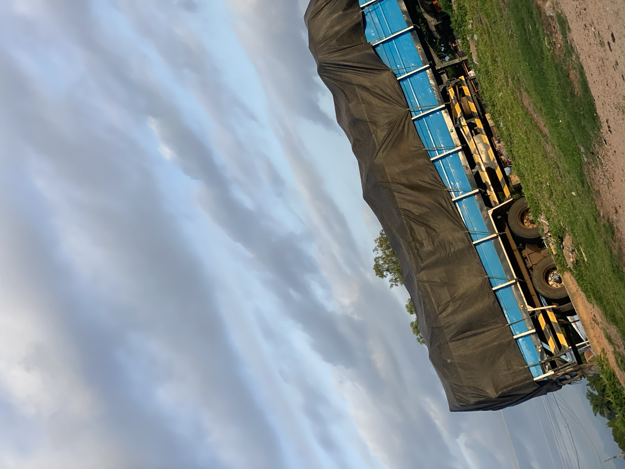 A large, blue truck covered with a black tarp is parked on a grassy incline. The truck is tilted to the left, with its wheels visible on the right side. The tarp creates a wavy silhouette over the cargo area. The background consists of a blue sky with scattered gray clouds. Trees and vegetation are visible on the left side of the image, and power lines can be seen in the distance. The ground is a mixture of dirt and grass. The scene appears to be in Jalingo, Nigeria, given the context.