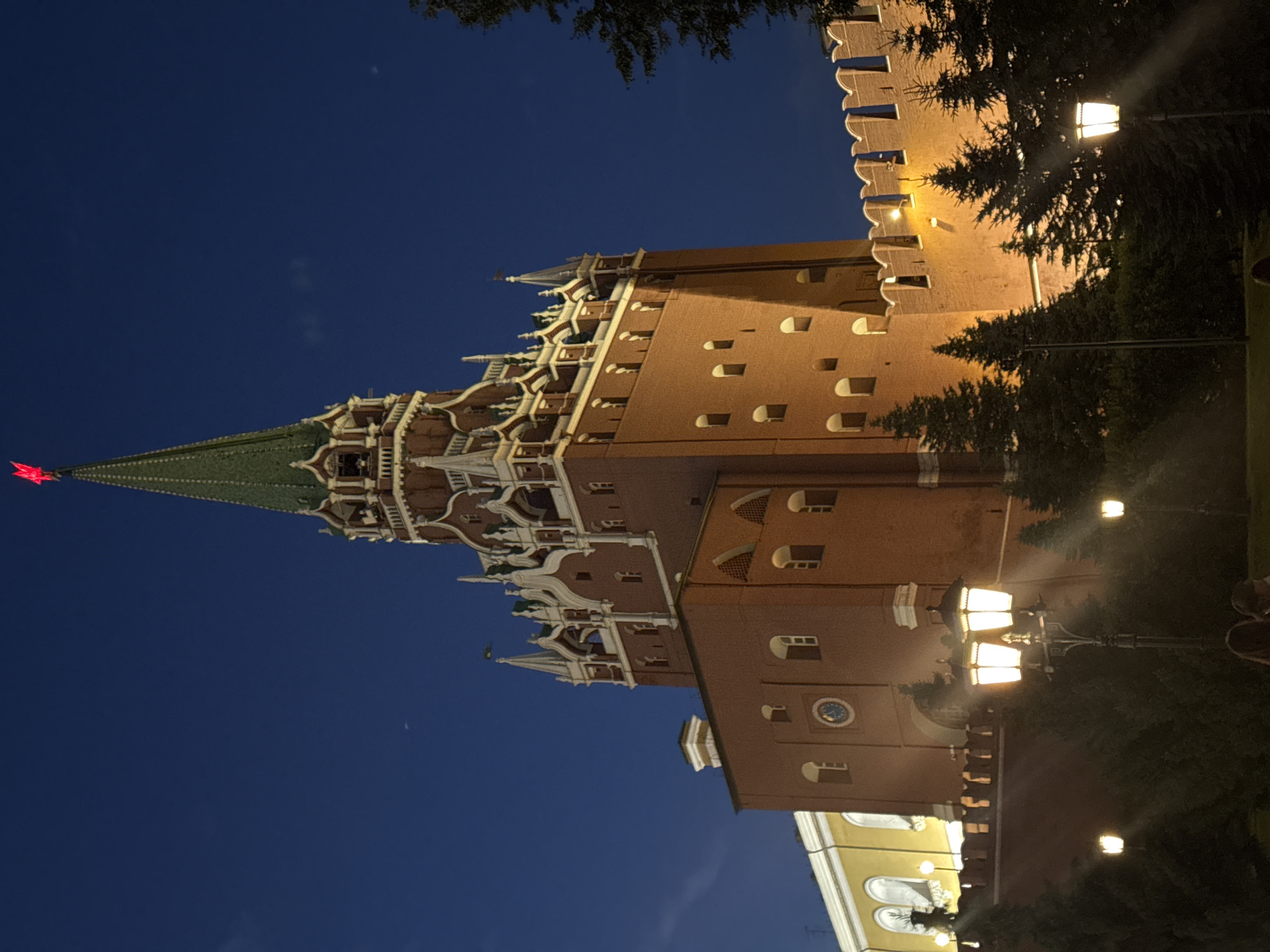 The image depicts the Spasskaya Tower of the Moscow Kremlin at night. The tower, constructed of red brick, is illuminated, highlighting its ornate white detailing and green spired roof, topped with a red star. The dark blue night sky serves as the backdrop. In the foreground, there are several illuminated street lamps casting a warm glow on the scene. The base of the tower is partially obscured by dark green evergreen trees. A low, crenelated brick wall, part of the Kremlin's fortifications, runs horizontally across the mid-ground, also lit from above. To the left of the tower, a section of a yellow building with arched windows is visible. The overall atmosphere is one of architectural grandeur and historical significance, captured during nighttime hours.
