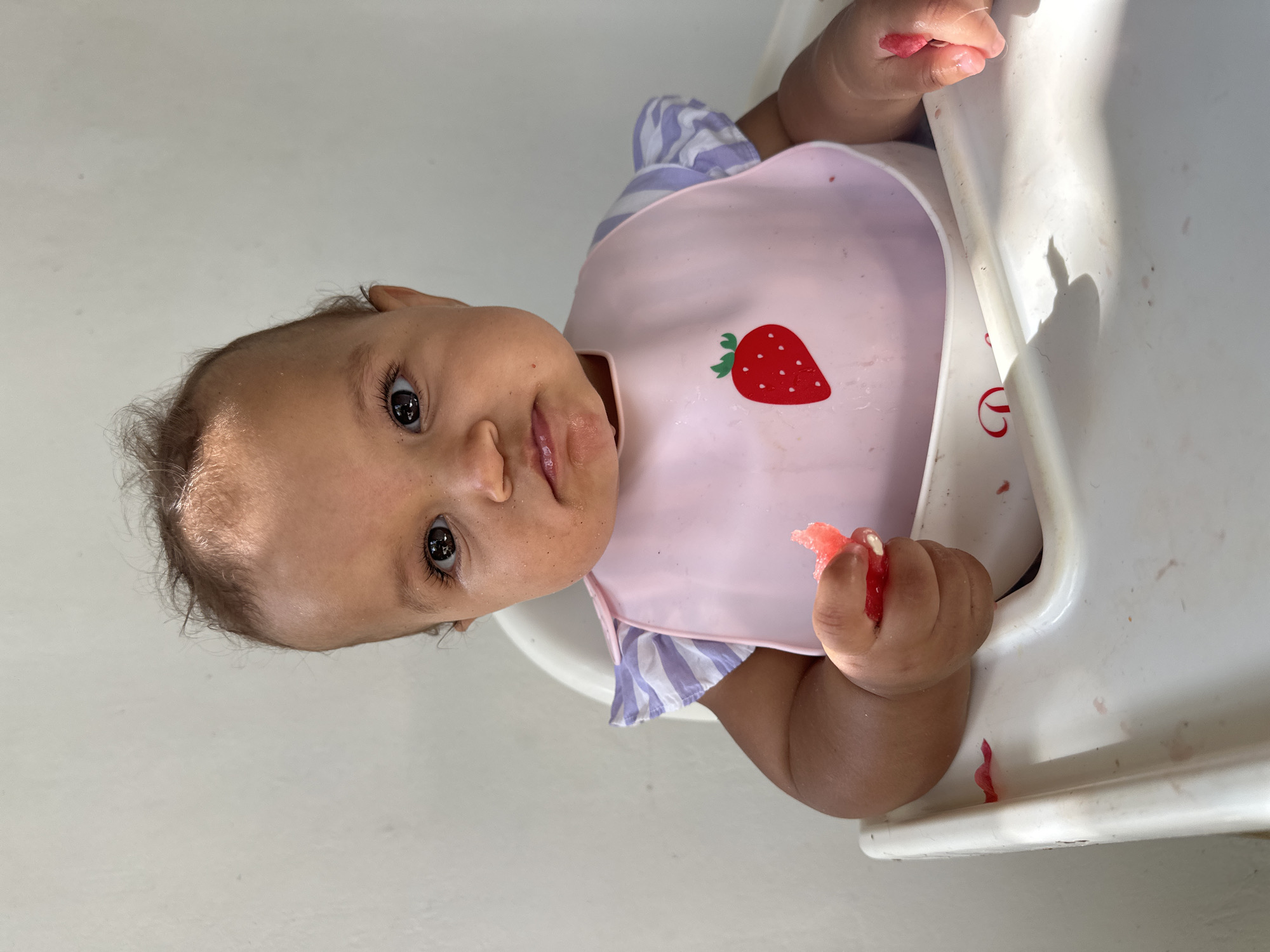 A baby sits in a white high chair, wearing a pink bib with a strawberry design. The baby is holding a piece of watermelon in its right hand and has watermelon juice around its mouth. The background is a plain white wall.