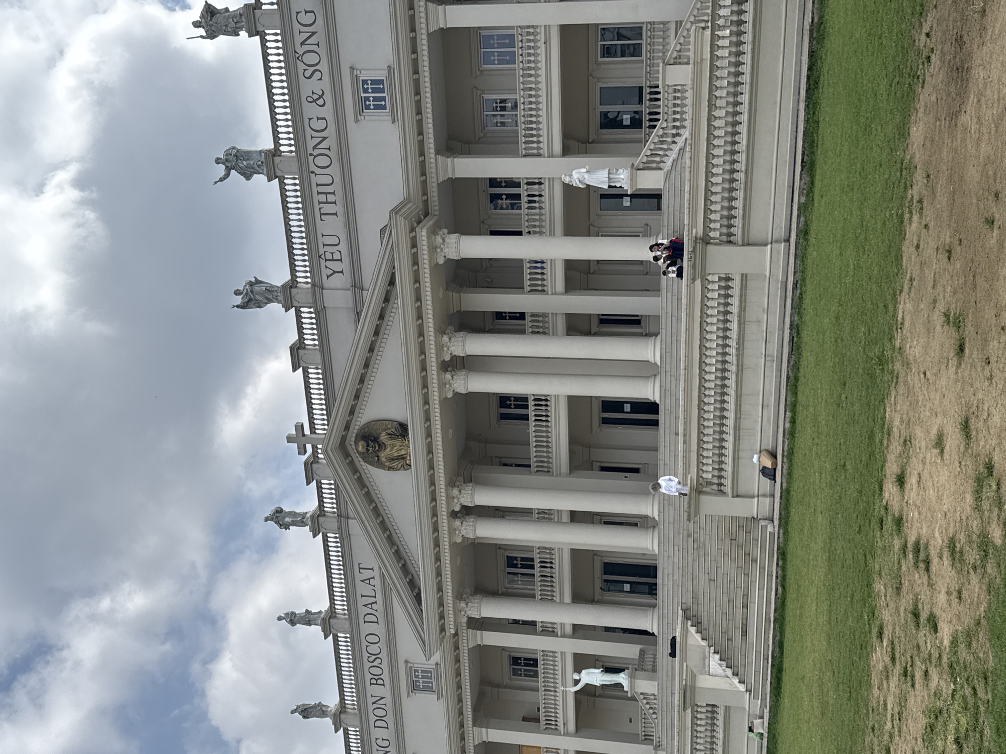 The image depicts a large, white building with classical architectural elements, possibly a church or religious institution. The facade features numerous columns, statues, and decorative railings. Inscribed on the building is the text "YÊU THƯƠNG & SỐNG" and "TRUNG DON BOSCO DALAT", suggesting the location is Da Lat, Vietnam.
Several people are visible on the steps of the building, some sitting and others standing. The environment includes a cloudy sky in the background and a grassy area to the side of the building. The scene appears to be a tourist location or a place of worship.