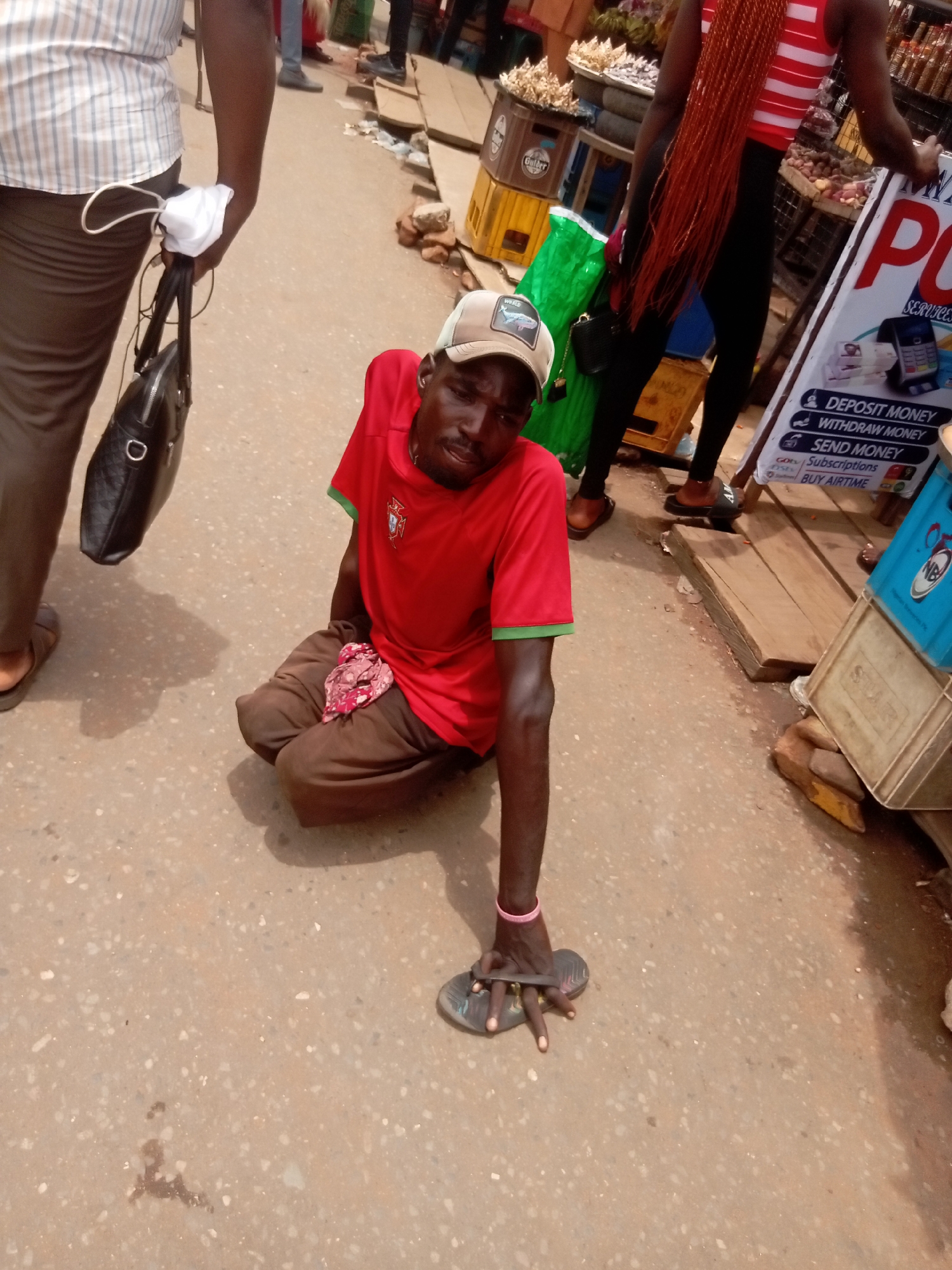 A man without legs is on the ground in Nkpor, Nigeria. He is wearing a red Portugal jersey and brown trousers. His left hand grips a flip-flop on the ground. A man holding a black bag and white face mask stands nearby. Other people and market stalls are visible in the background.