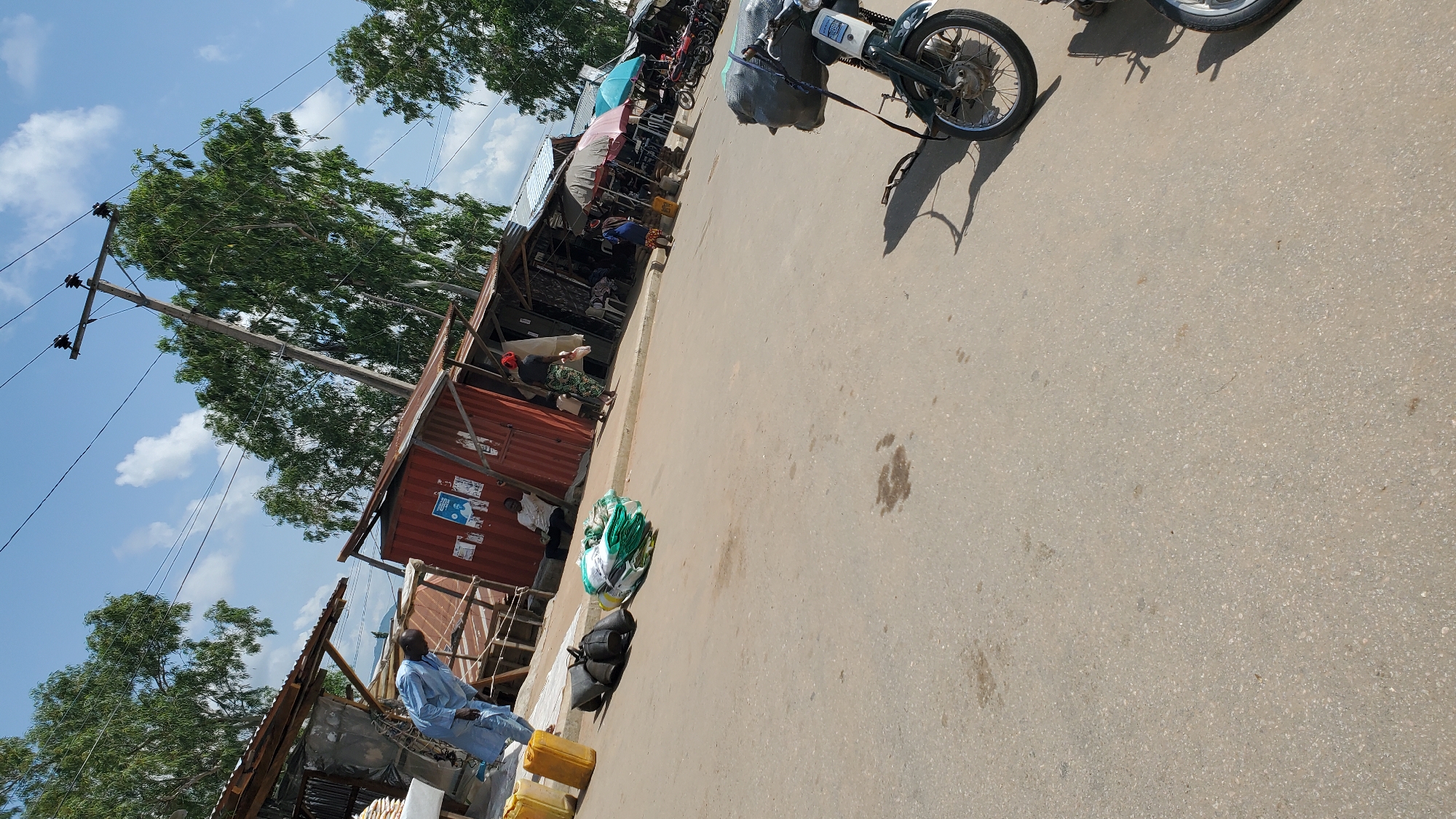 In Jalingo, Nigeria, a street scene unfolds under a bright, cloudy sky. The image captures a bustling market or roadside area with several structures, likely makeshift shops, lining the paved road. In the foreground, a parked motorcycle is visible, its front wheel and part of its frame in focus. Further down the road, other motorcycles are parked in a row.
Several people are present. A man in a light blue traditional garment is bent over near some yellow canisters and what appear to be bags of goods on the sidewalk. Another man sits on a step near a red shipping container structure. A woman wearing a headscarf and a patterned skirt walks away from the camera, carrying a white object. Another woman in blue stands near some umbrellas. The structures themselves are varied, including some made of corrugated metal and wood, and one appears to be a repurposed shipping container.
The environment suggests an outdoor market or a commercial street. Overhead, power lines stretch across the frame, supported by a utility pole. Trees with lush green foliage are visible behind the structures, contributing to a slightly rural or peri-urban feel. The road surface is asphalt, with some minor stains and textures visible. The overall impression is of a lively, everyday scene in a Nigerian town.