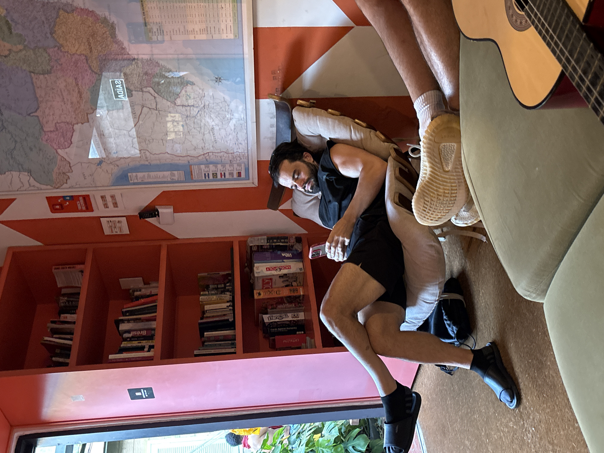 This image captures a relaxed indoor common area, possibly a hostel or lounge, located in São Paulo, Brazil. The scene features two men in a casual setting. One man, with a beard and dark hair, is resting comfortably on a light-colored armchair, wearing a dark sleeveless top and shorts. He appears to be holding a small white object, possibly a phone or remote. The second person is partially visible, showing their legs. One leg, clad in light shorts, white socks, and light-colored sneakers with "BOOST" visible on the sole, is propped up. The other leg, in dark shorts, black socks, and black Adidas slides, rests on the floor. An acoustic guitar is visible next to this individual.
The room's decor includes walls painted with a distinctive orange and white striped geometric pattern. A large framed map of South America is prominently displayed on the wall, showing regions and countries like Bolivia, Paraguay, and Argentina, with some text like "ADIVIA" (reversed) visible due to reflections. Below the map, an orange and pink bookshelf unit is stocked with numerous books, including one with "BRASIL" on its spine, and a collection of board games such as Monopoly, Vida, Dixit, Trouble, Catan, Risk, Carcassonne, and Earth Against Humanity. A small red sign on the wall clearly states "TROCA DE LIVROS" (Book Exchange in Portuguese). A black backpack or bag is on the carpeted floor, which is light brown. Through an opening at the bottom of the frame, green plants are visible, suggesting either an outdoor view or indoor foliage. The overall atmosphere is one of leisure and communal activity.